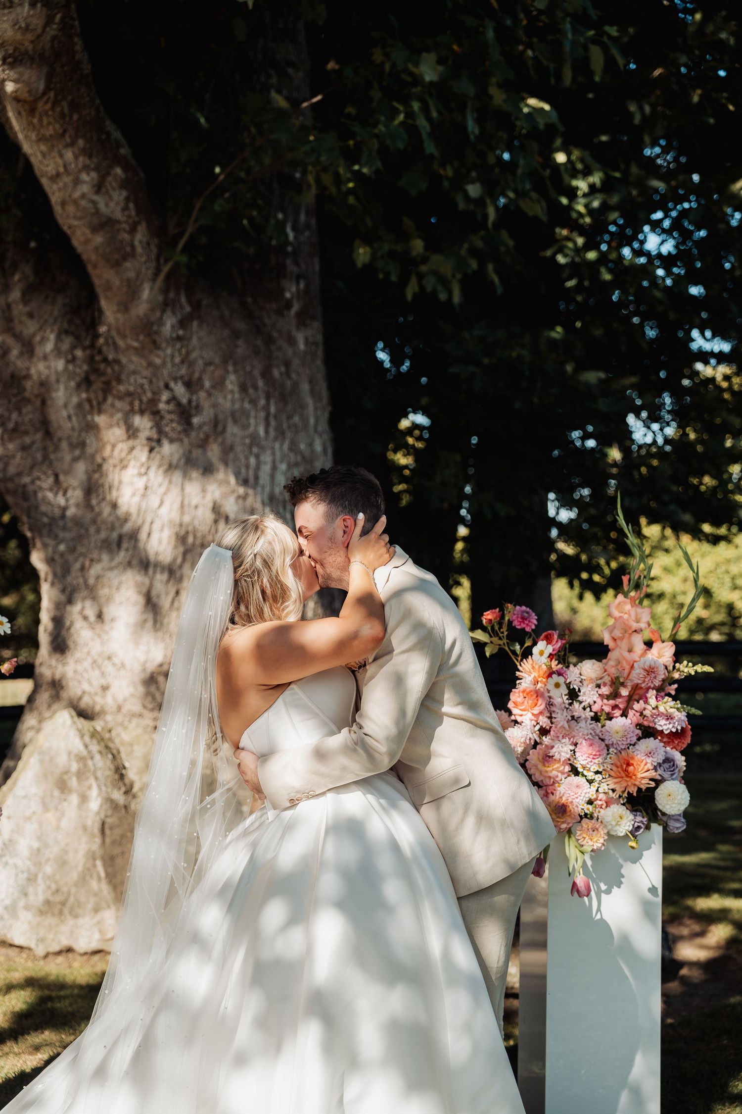 Bride and groom sharing first kiss at outdoor wedding ceremony beside floral arrangement.