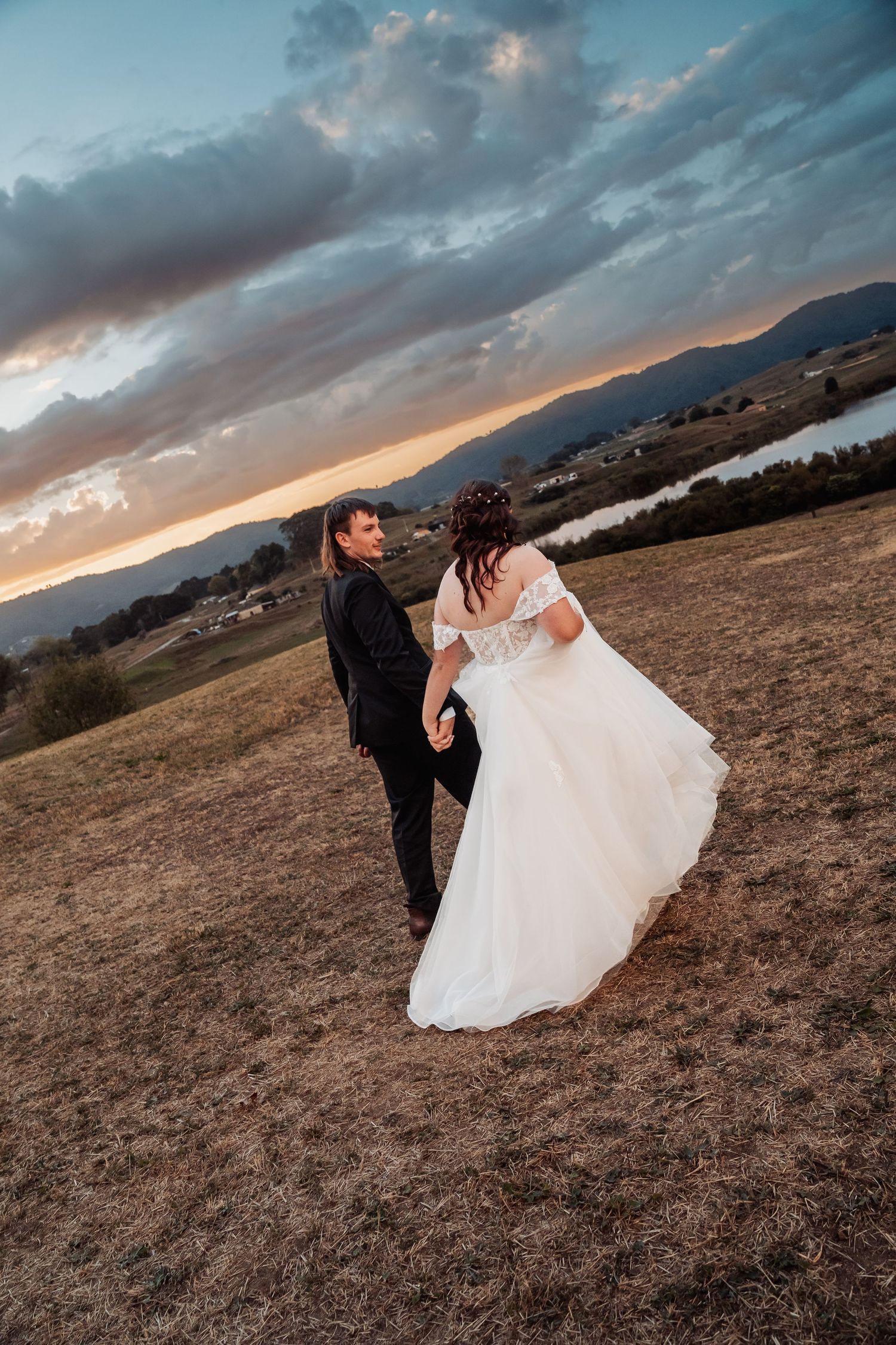 Bride and groom walking hand in hand across an open field at dramatic sunset with mountains.