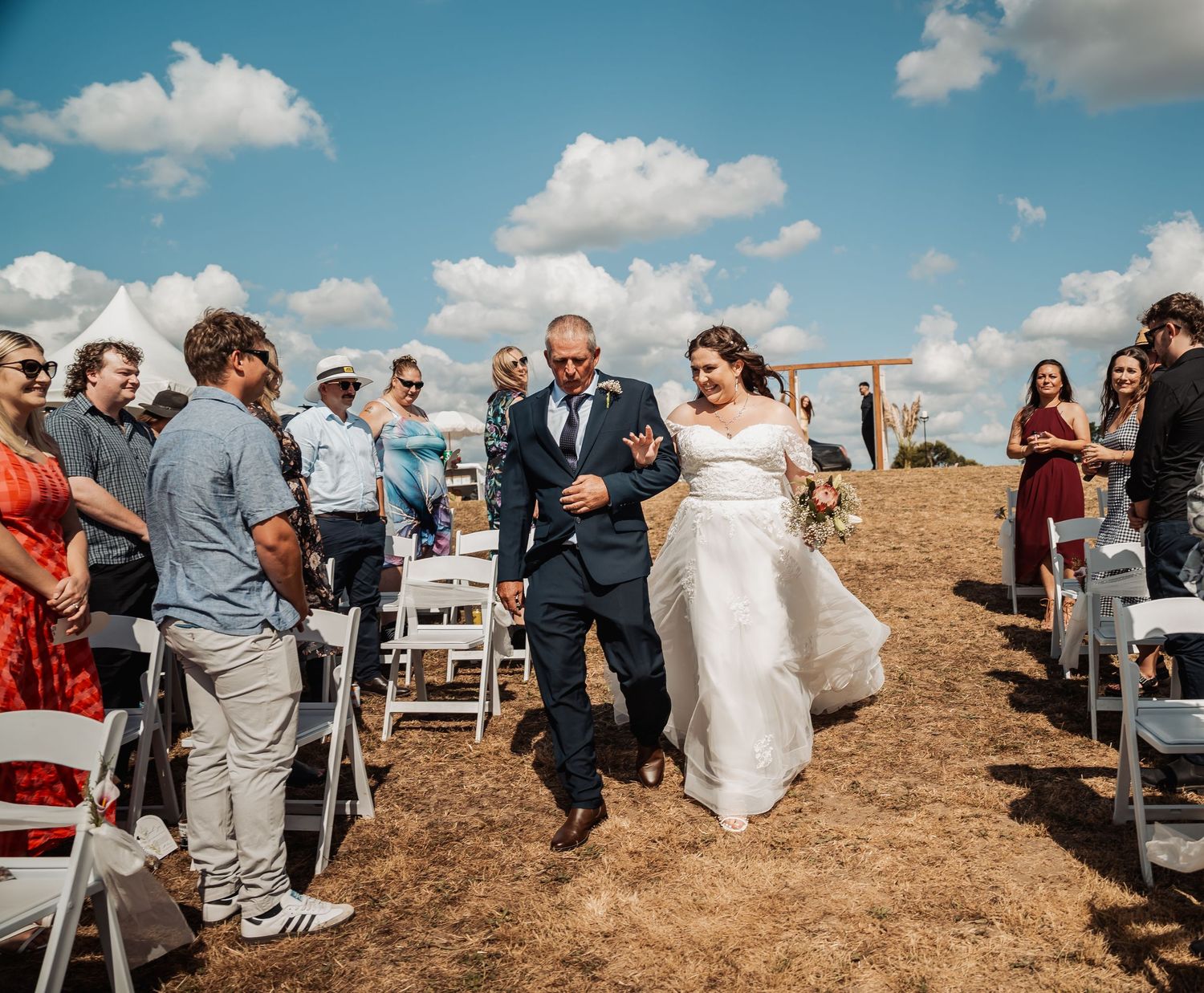 Father walks bride down outdoor aisle surrounded by guests under blue sky on dry grassy field.