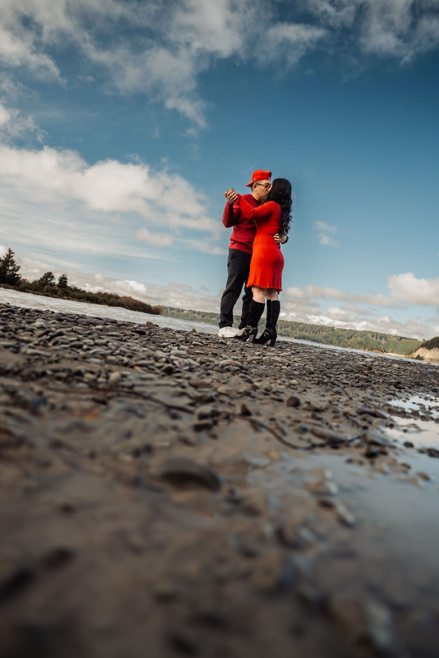 Couple in red outfits embrace and kiss on a rocky shoreline under a blue cloudy sky.