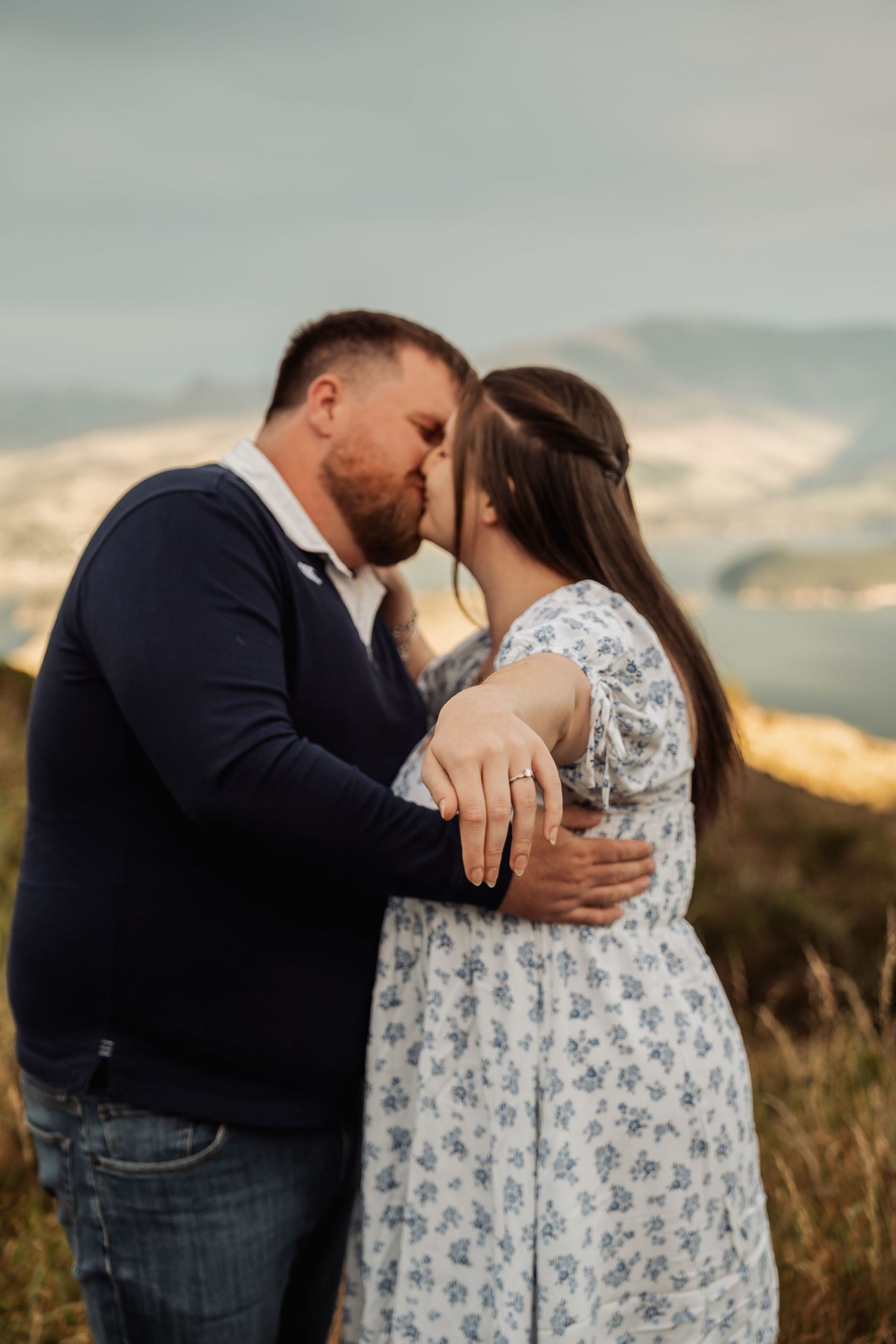 Couple sharing a romantic kiss outdoors at sunset, woman showing engagement ring, scenic mountain view background.