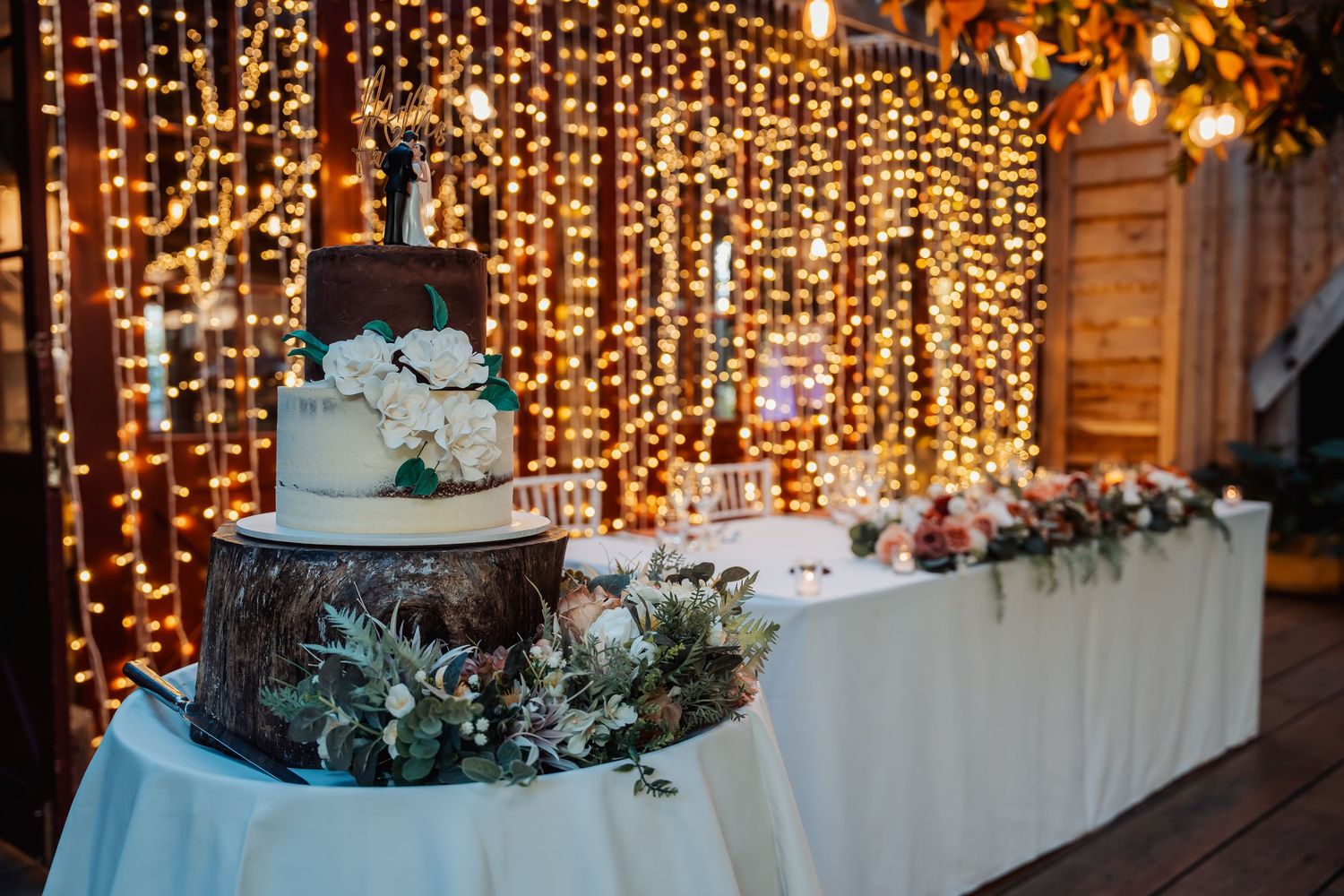 Two-tier white wedding cake on a wood stump stand, decorated with flowers, set against a warm fairy light backdrop.