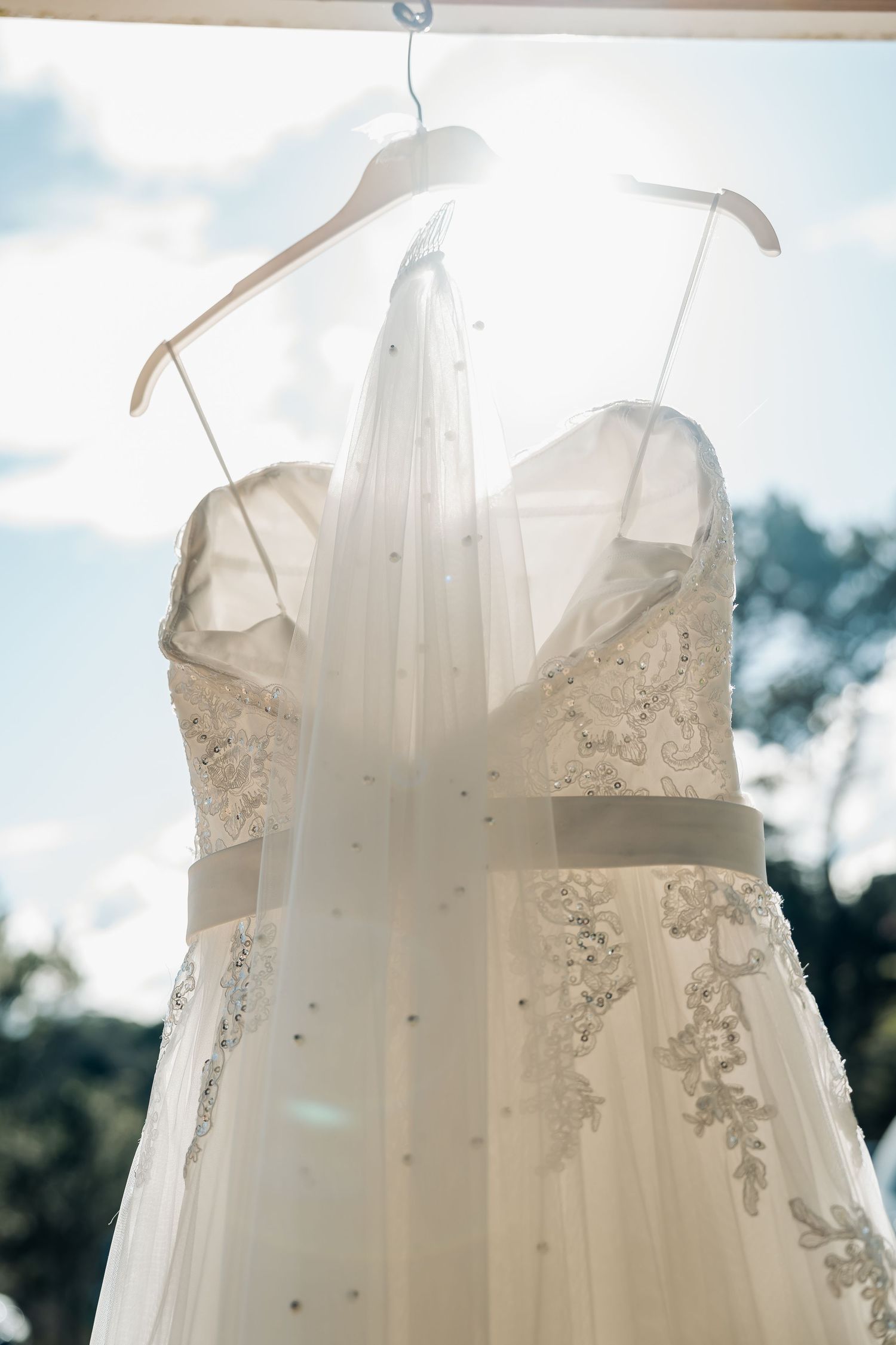 A white wedding dress with delicate lace details hangs on display in soft natural lighting against a blurred outdoor backdrop.