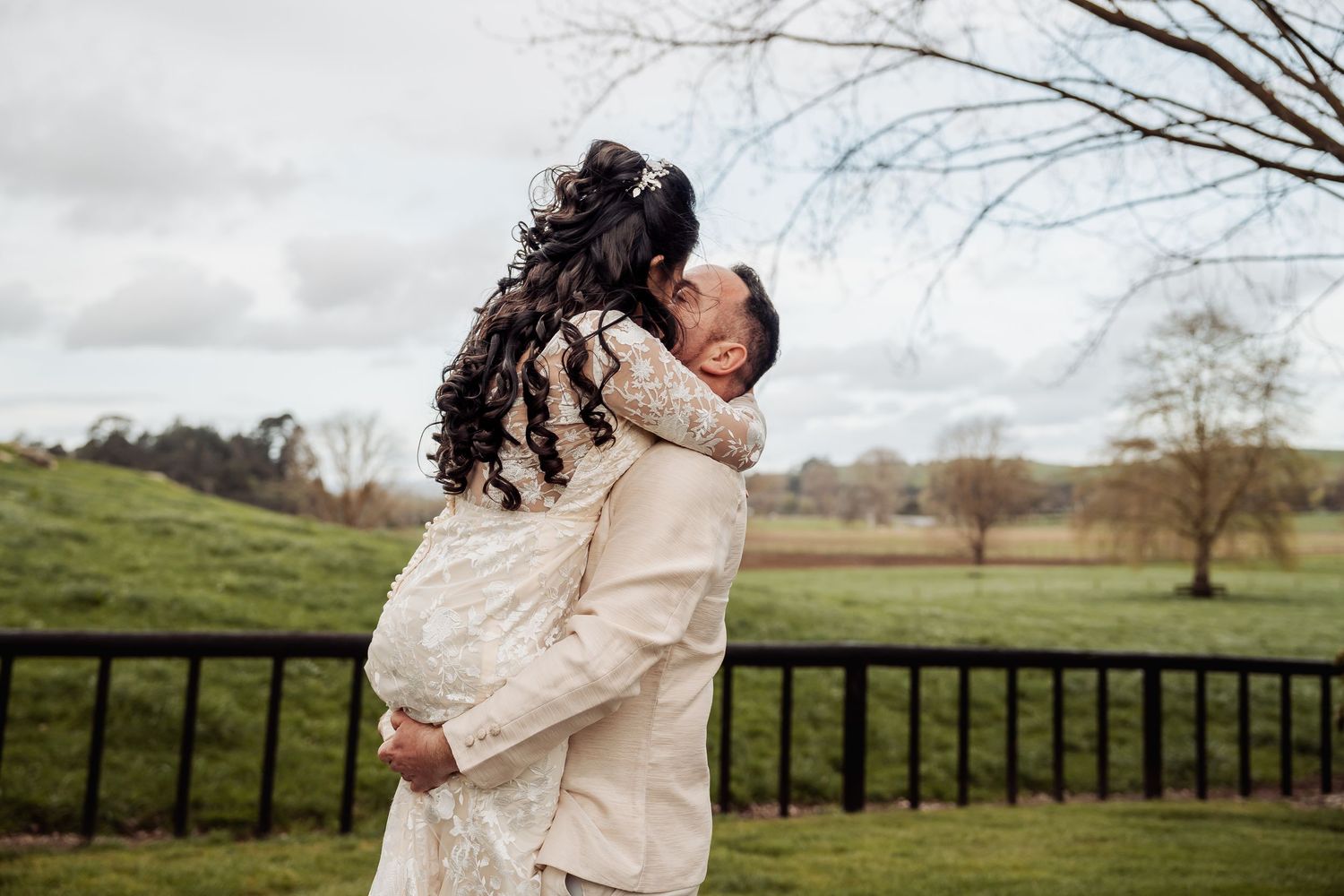 A couple embraces on a gravel path surrounded by bare trees and green pastures.