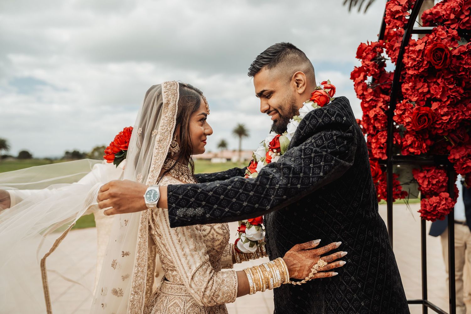 Romantic outdoor photoshoot with red floral decorations against a cloudy sky.
