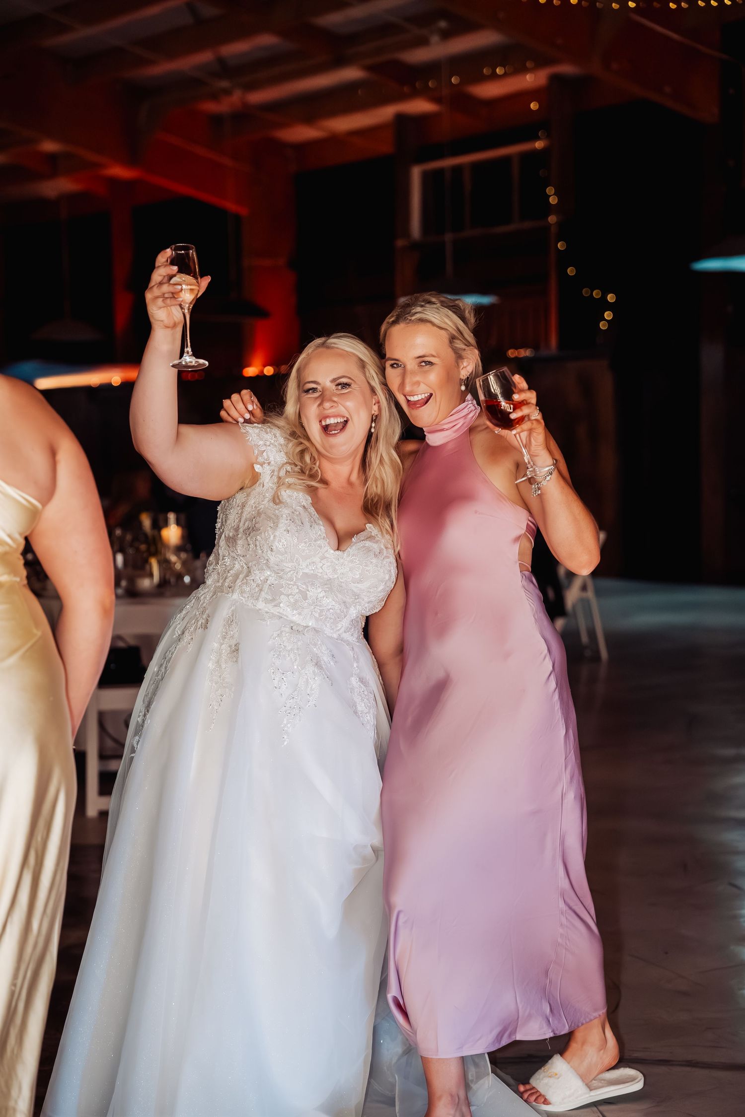 Joyful bride in lace gown and guest in pink dress raise wine glasses celebrating at reception.