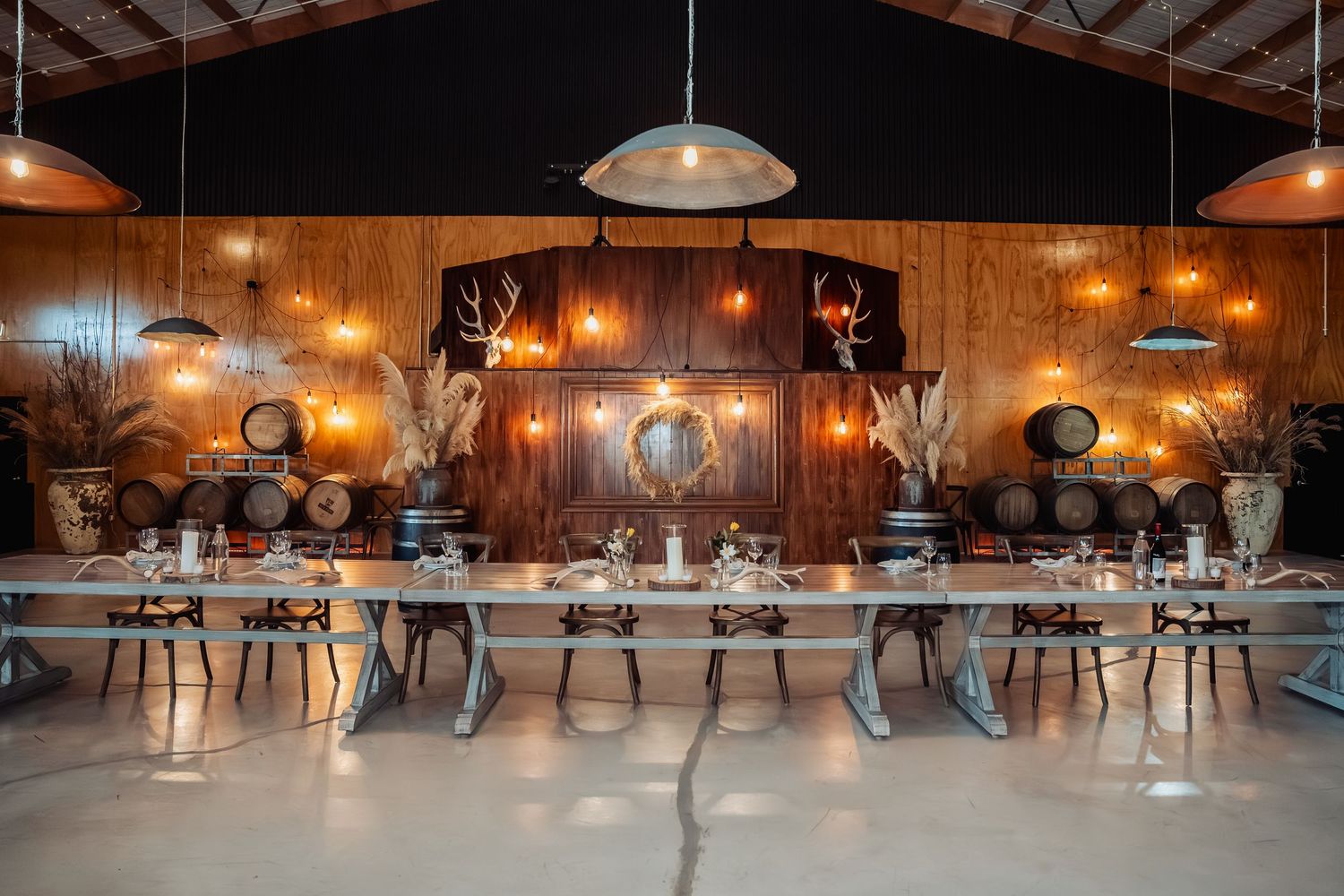 Rustic winery event space with long dining table, barrel decor, warm pendant lights, and antler mounts on wood-paneled walls.