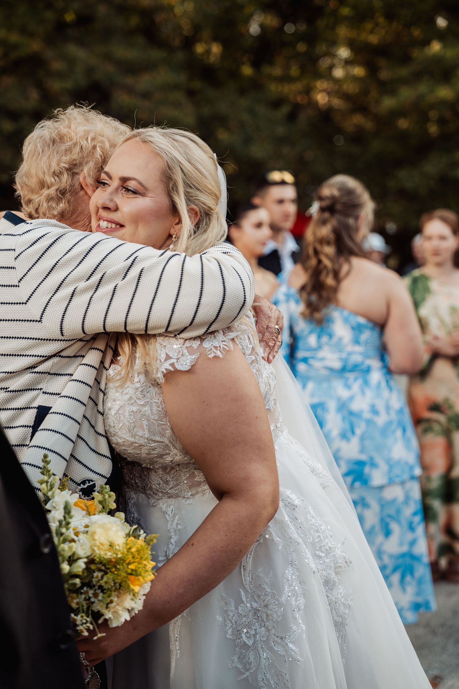 Bride in lace wedding gown hugging a guest while holding yellow bouquet at outdoor wedding reception.