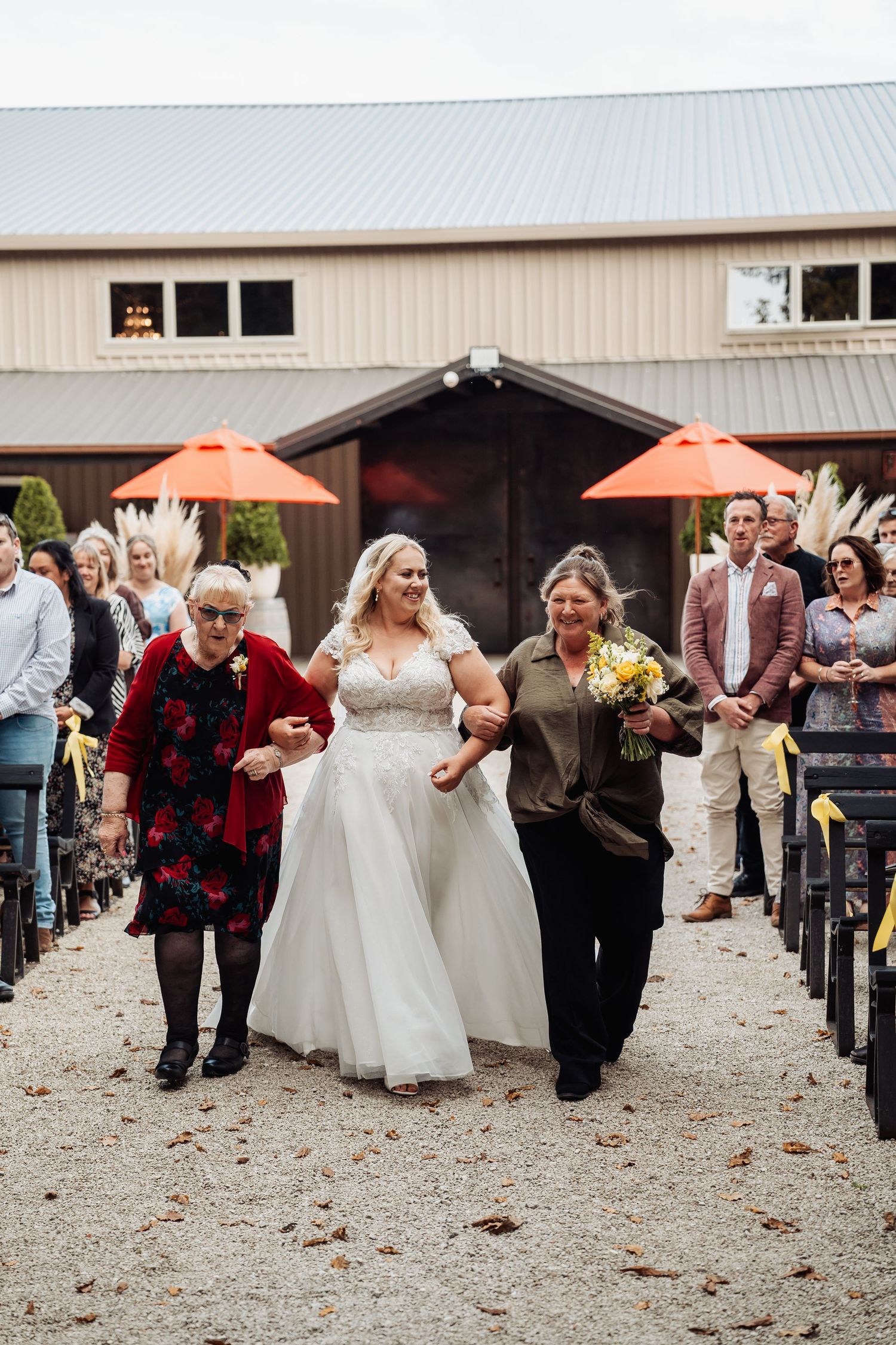 Bride walks down gravel aisle escorted by two women at outdoor barn wedding ceremony.