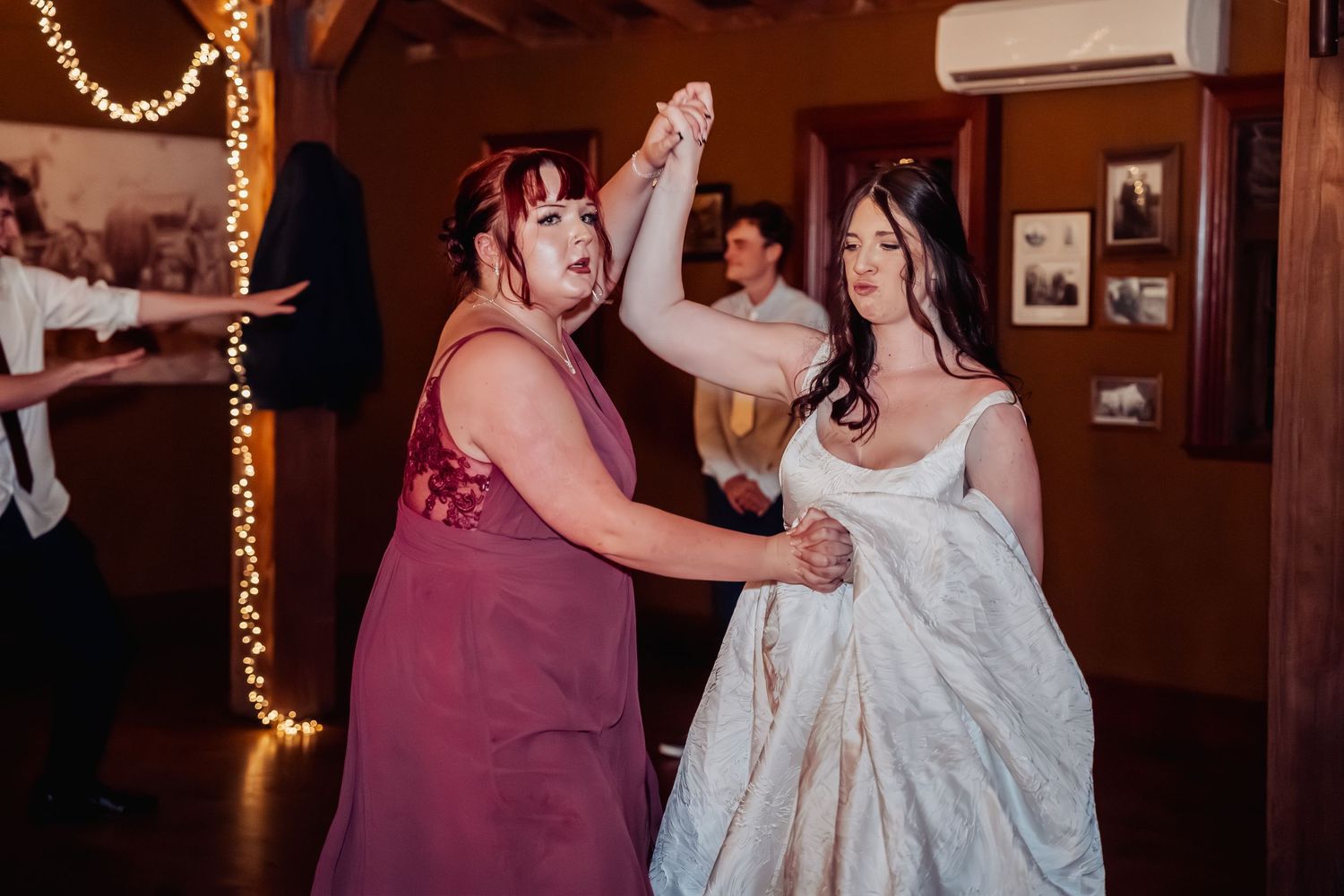Two women dancing joyfully at a wedding reception, one in a white bridal gown and one in a burgundy dress.