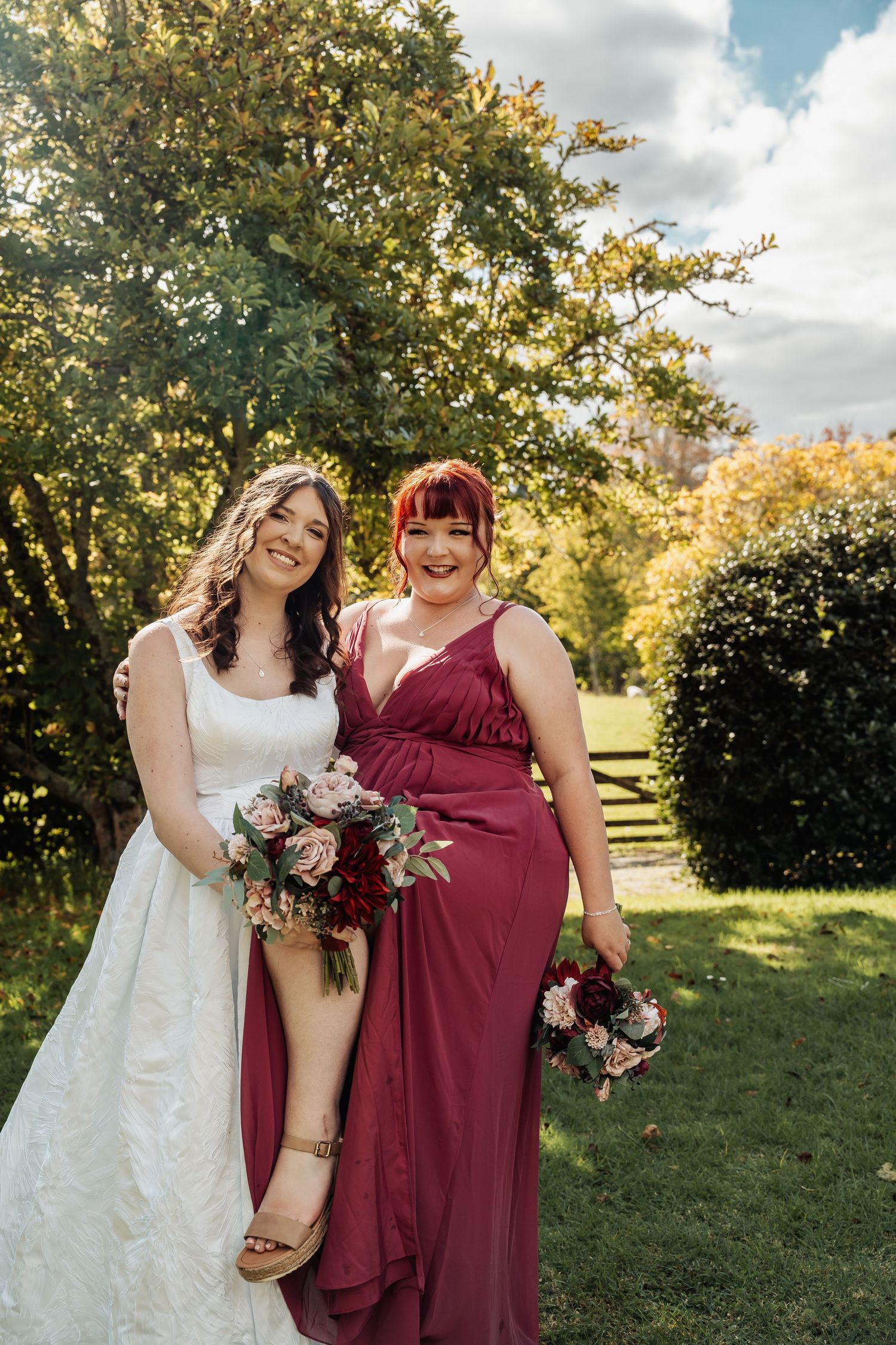 Bride in white gown and bridesmaid in burgundy dress holding bouquets outdoors at a wedding.