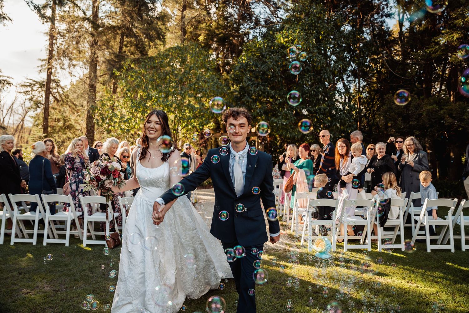 Newlyweds walk through bubble send-off past seated wedding guests in outdoor ceremony.