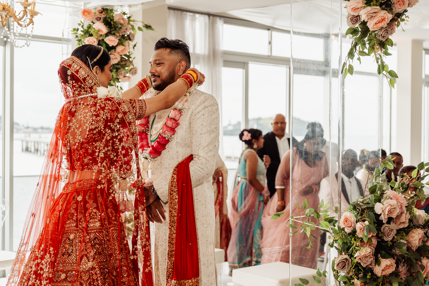 A wedding couple performs traditional Indian marriage rituals in an indoor venue with floral arrangements.