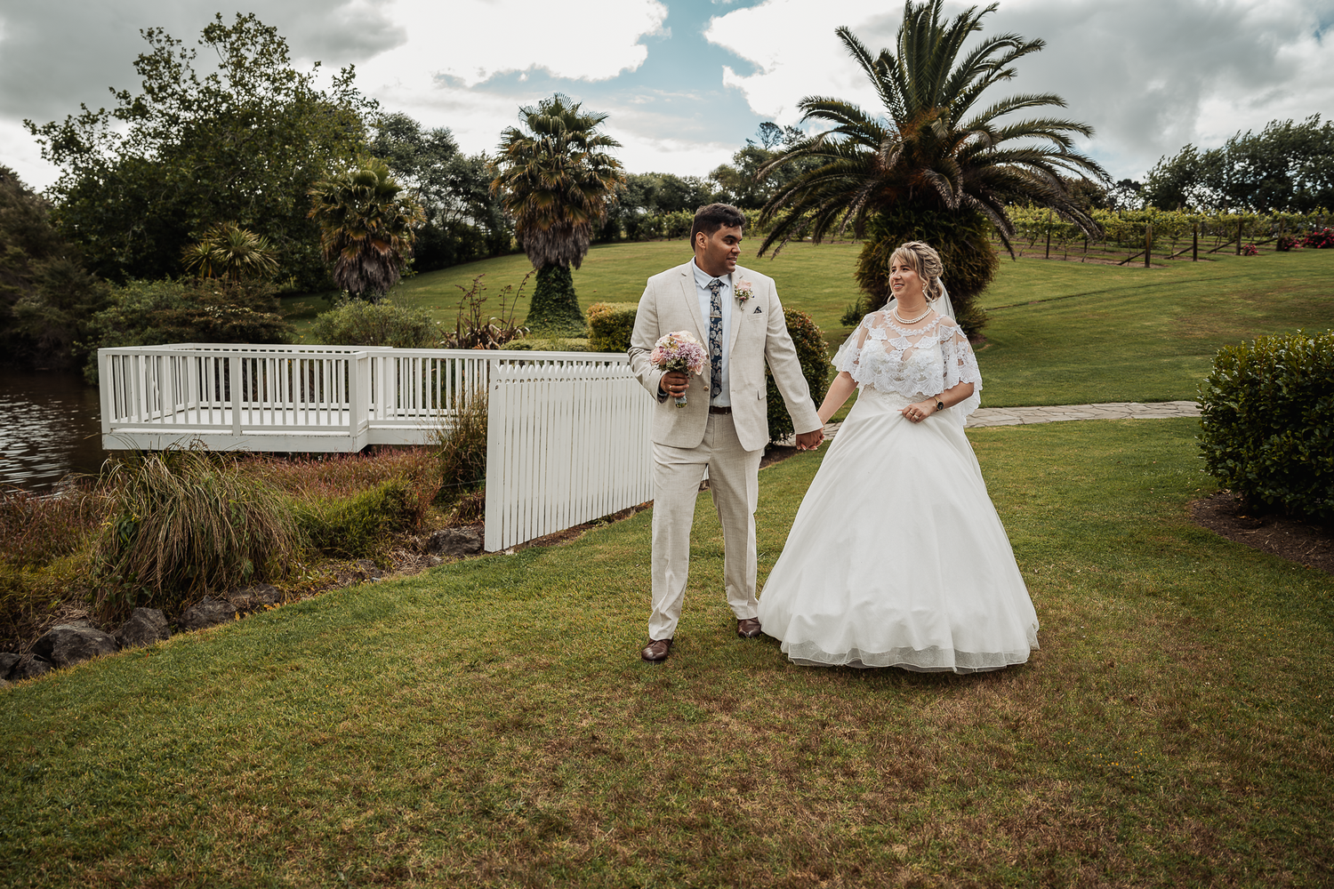 A couple walks along a garden path wearing elegant wedding attire in white.