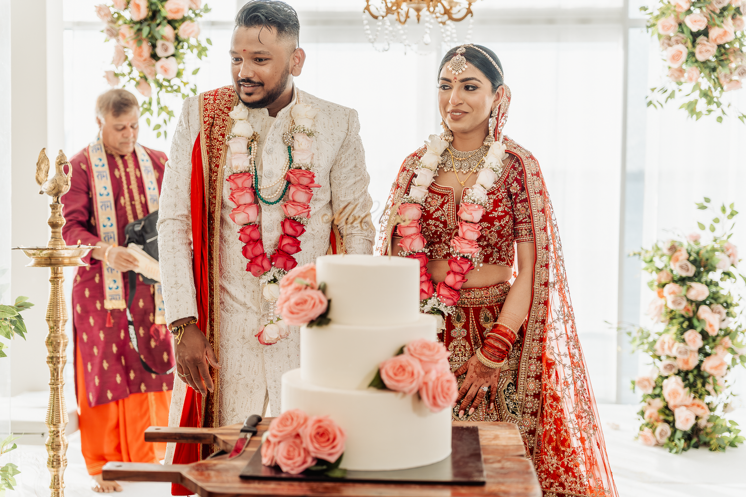 A wedding couple stands beside their white tiered cake decorated with pink flowers.