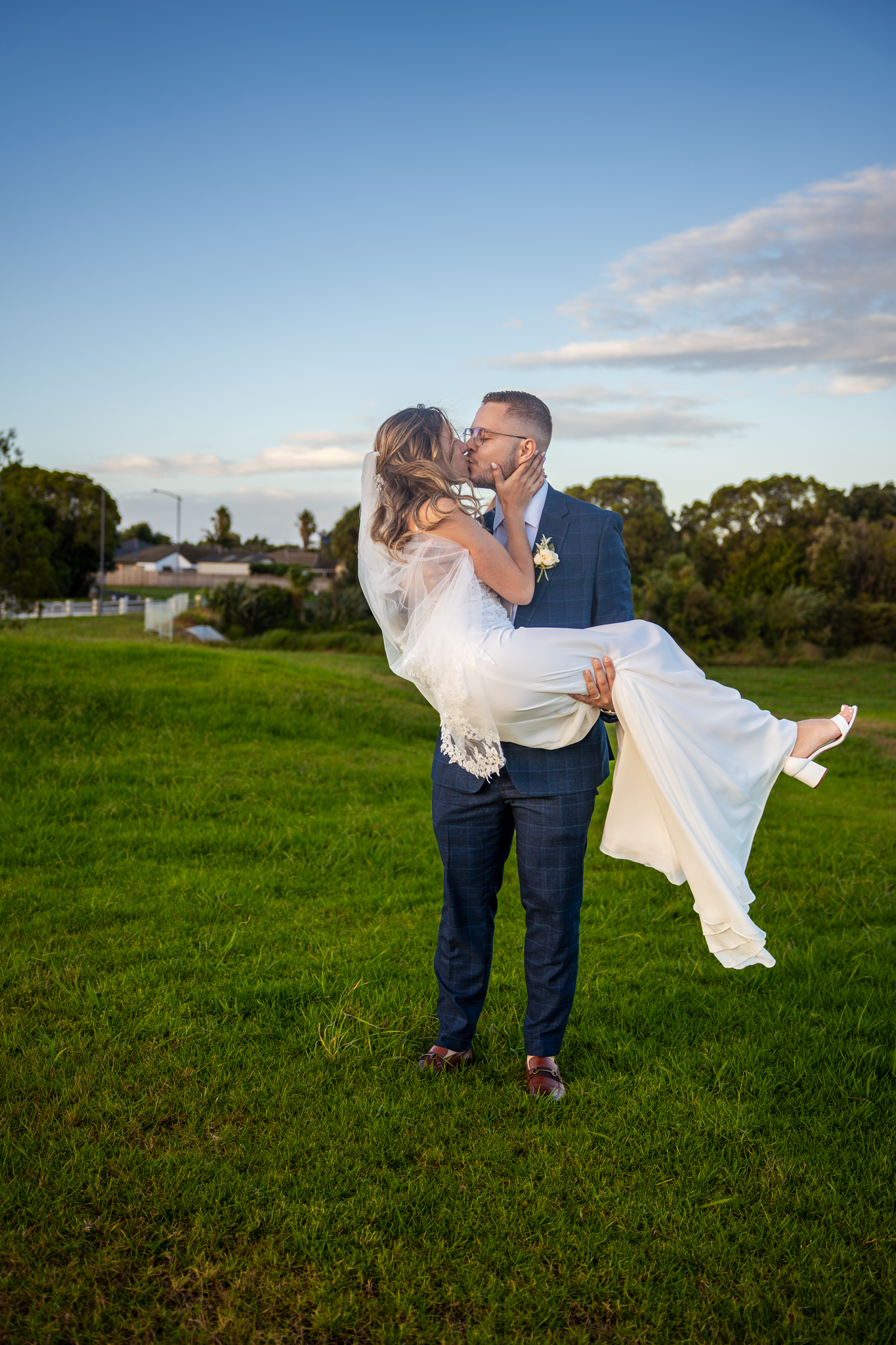 Groom carries bride in white dress during sunset on grassy field.