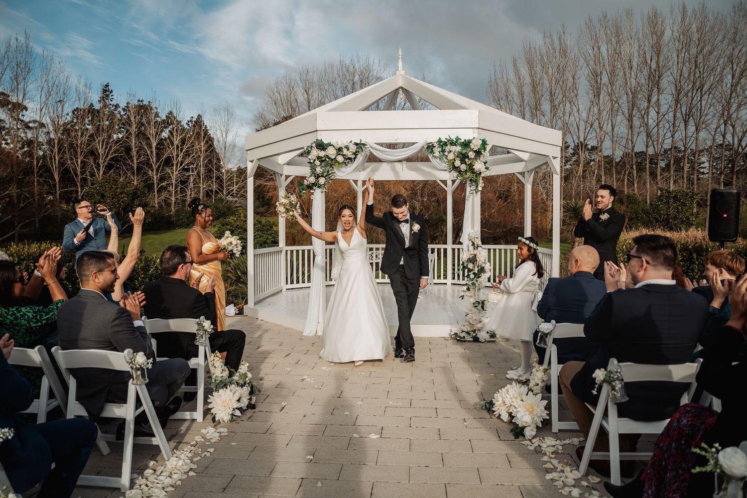 White wedding gazebo decorated with greenery set up for an outdoor ceremony with rows of chairs and guests.