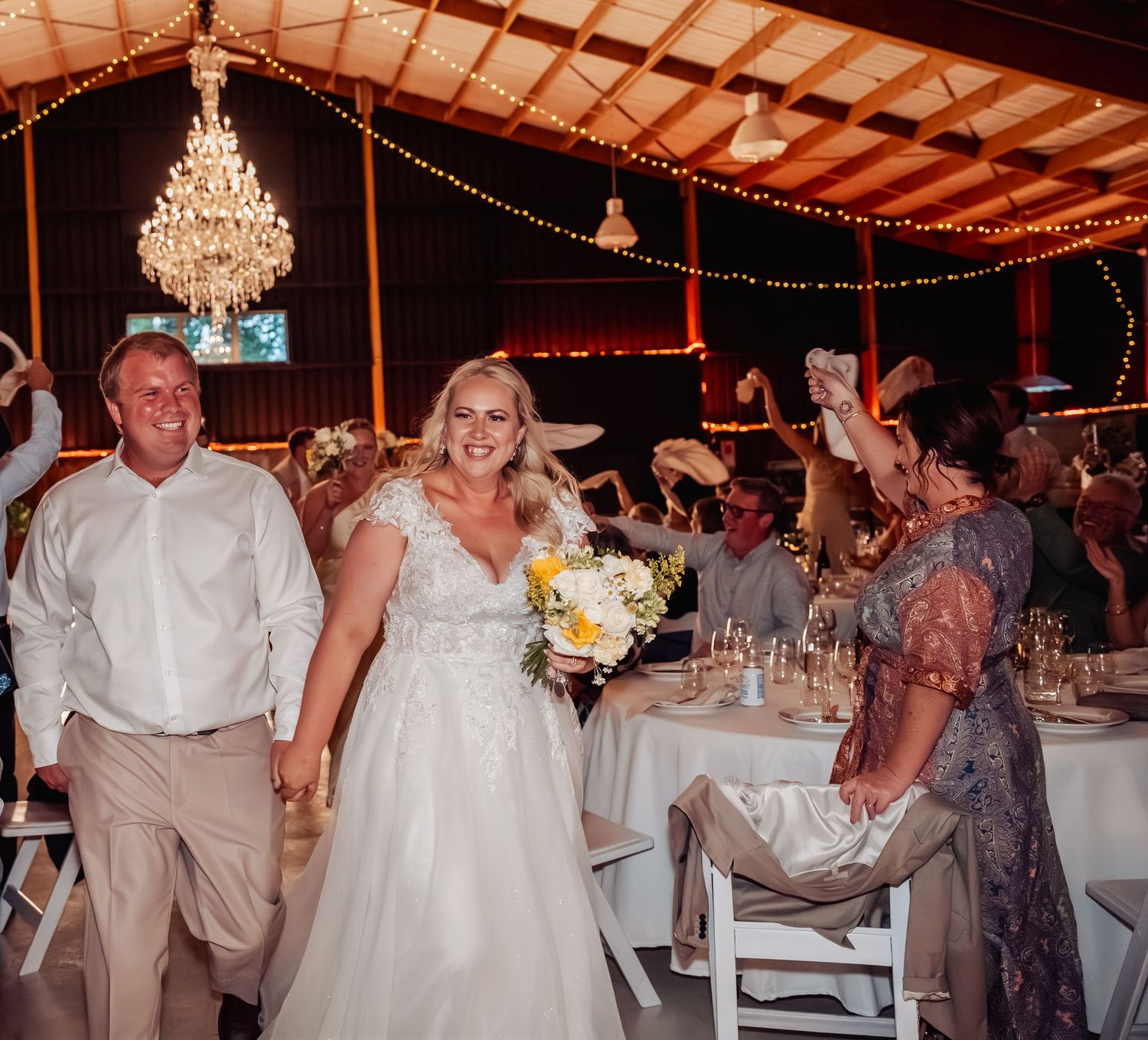 Newlyweds enter wedding reception in decorated barn with chandelier as guests cheer and wave napkins.