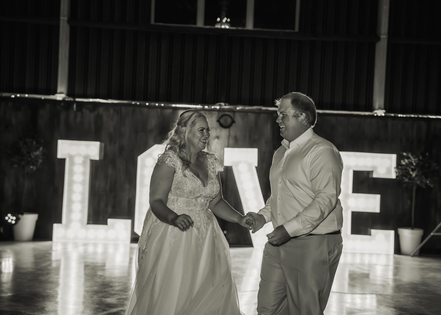 Black and white photo of bride and groom dancing before illuminated LOVE sign at reception.