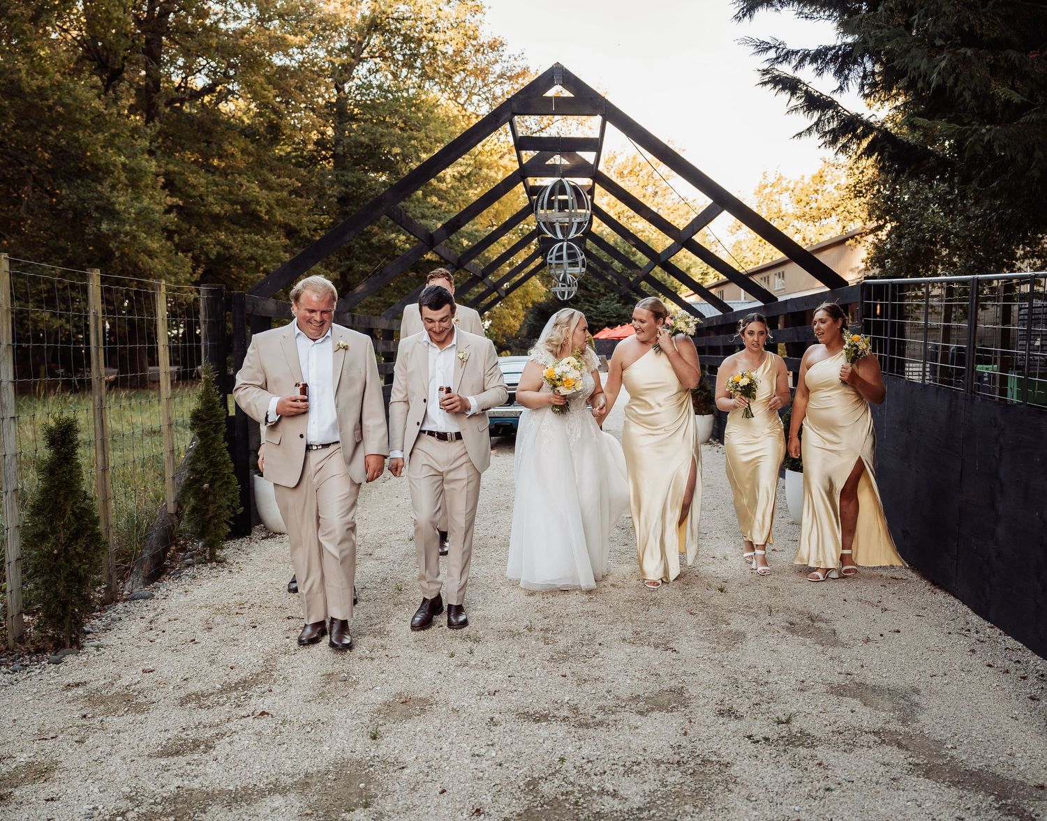 Wedding party walks together in elegant neutral attire at rustic outdoor venue.