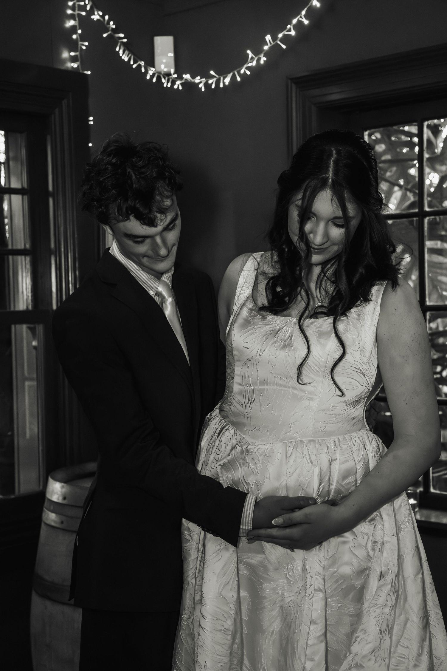 Black and white photo of pregnant bride and groom tenderly holding her baby bump indoors.