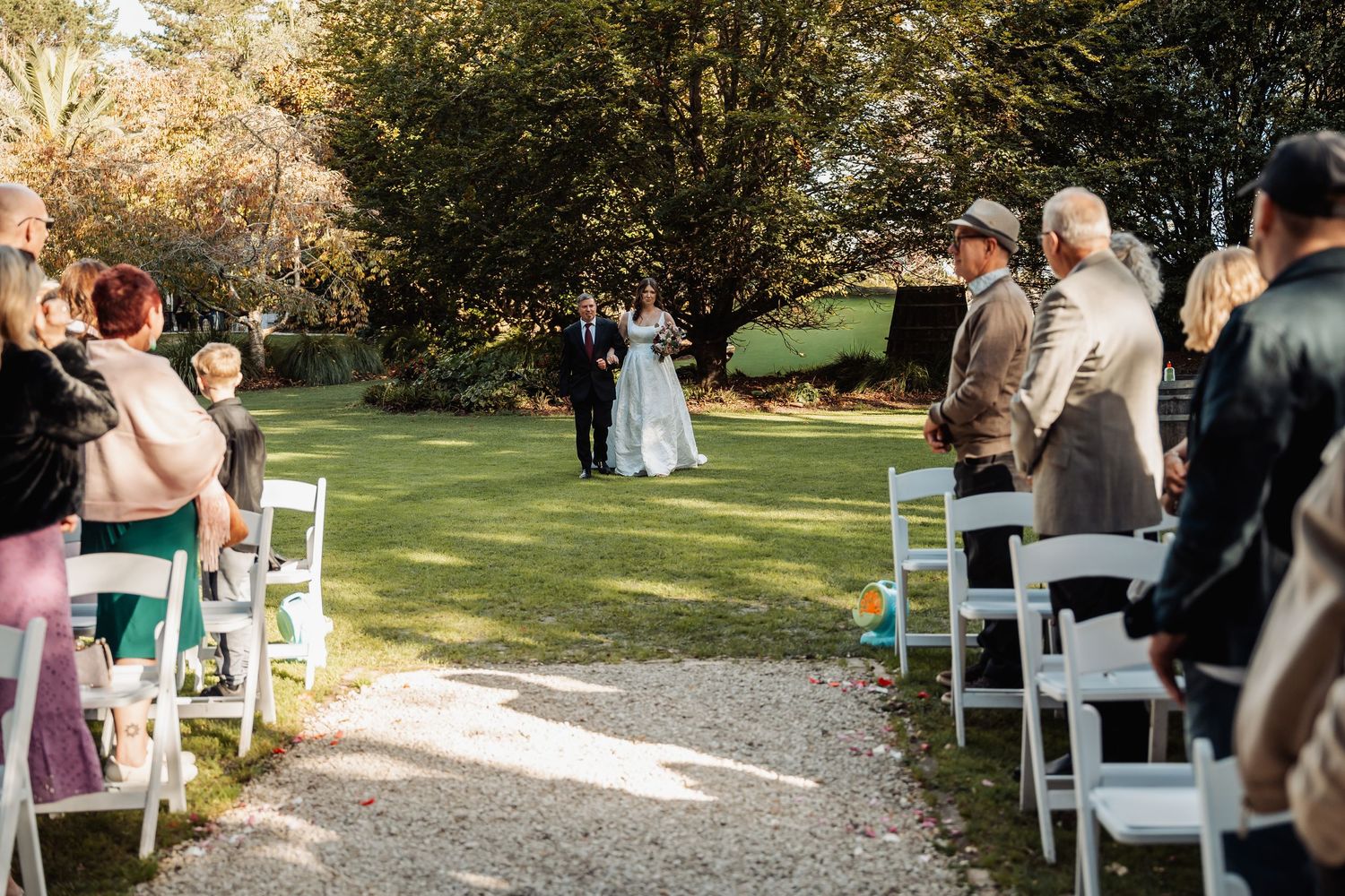 Bride and groom walk down outdoor wedding aisle surrounded by seated guests in sunny garden.