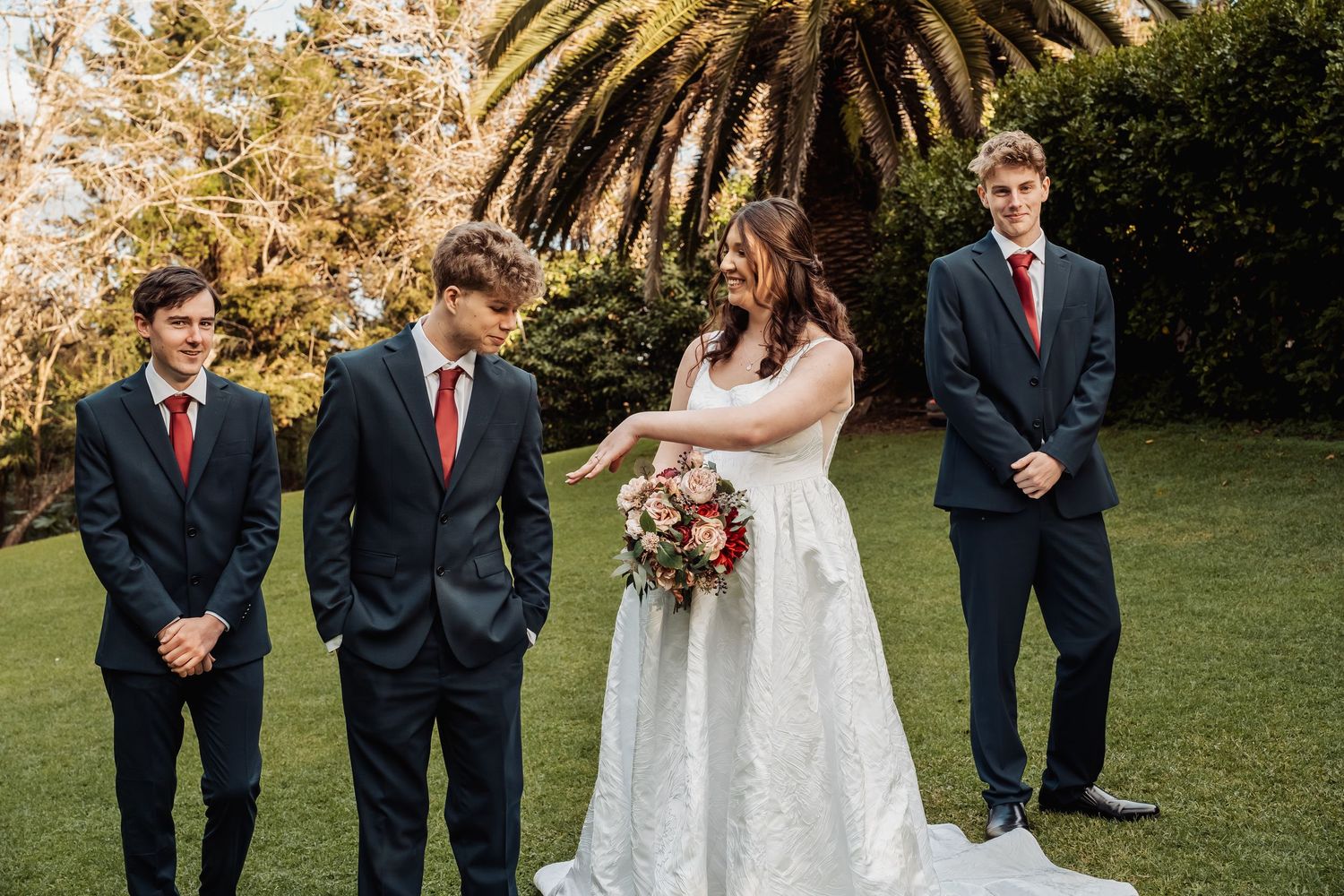 Bride in white gown holding bouquet with three groomsmen in navy suits on lawn.