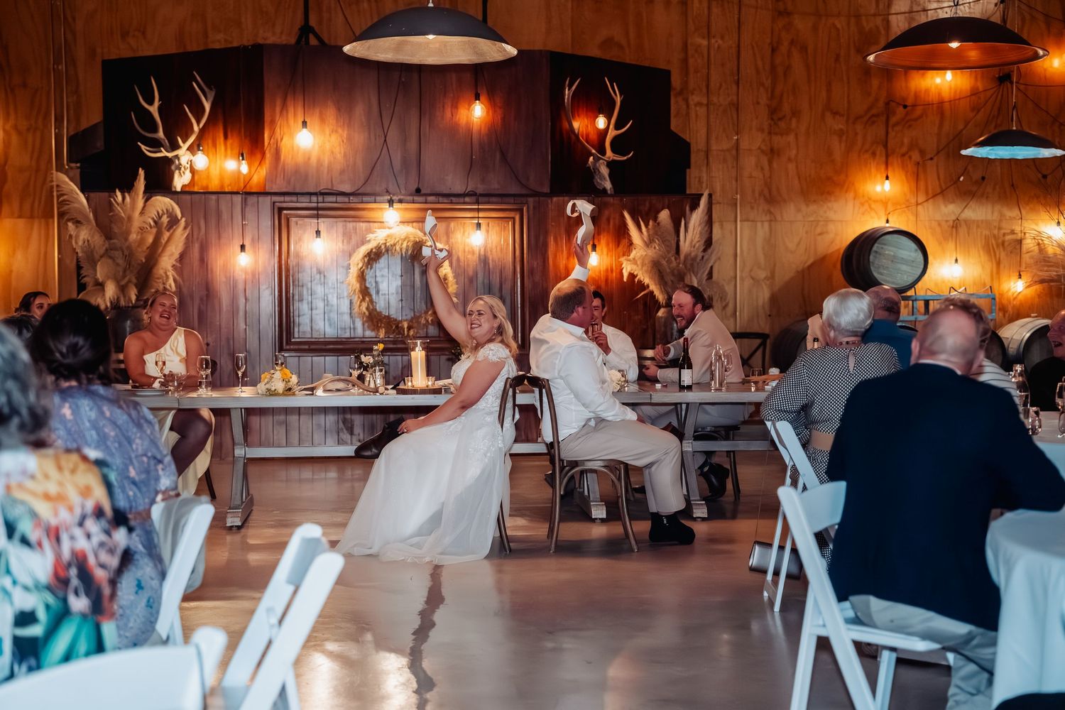 Two brides share a dance in a rustic wood-paneled wedding venue adorned with antler mounts and warm Edison bulb lighting.