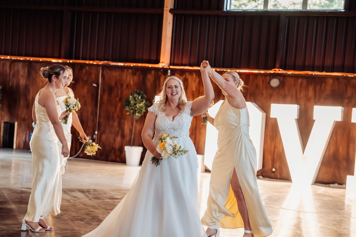 Bride dancing with bridesmaid in white gowns at rustic barn wedding reception with marquee letters.