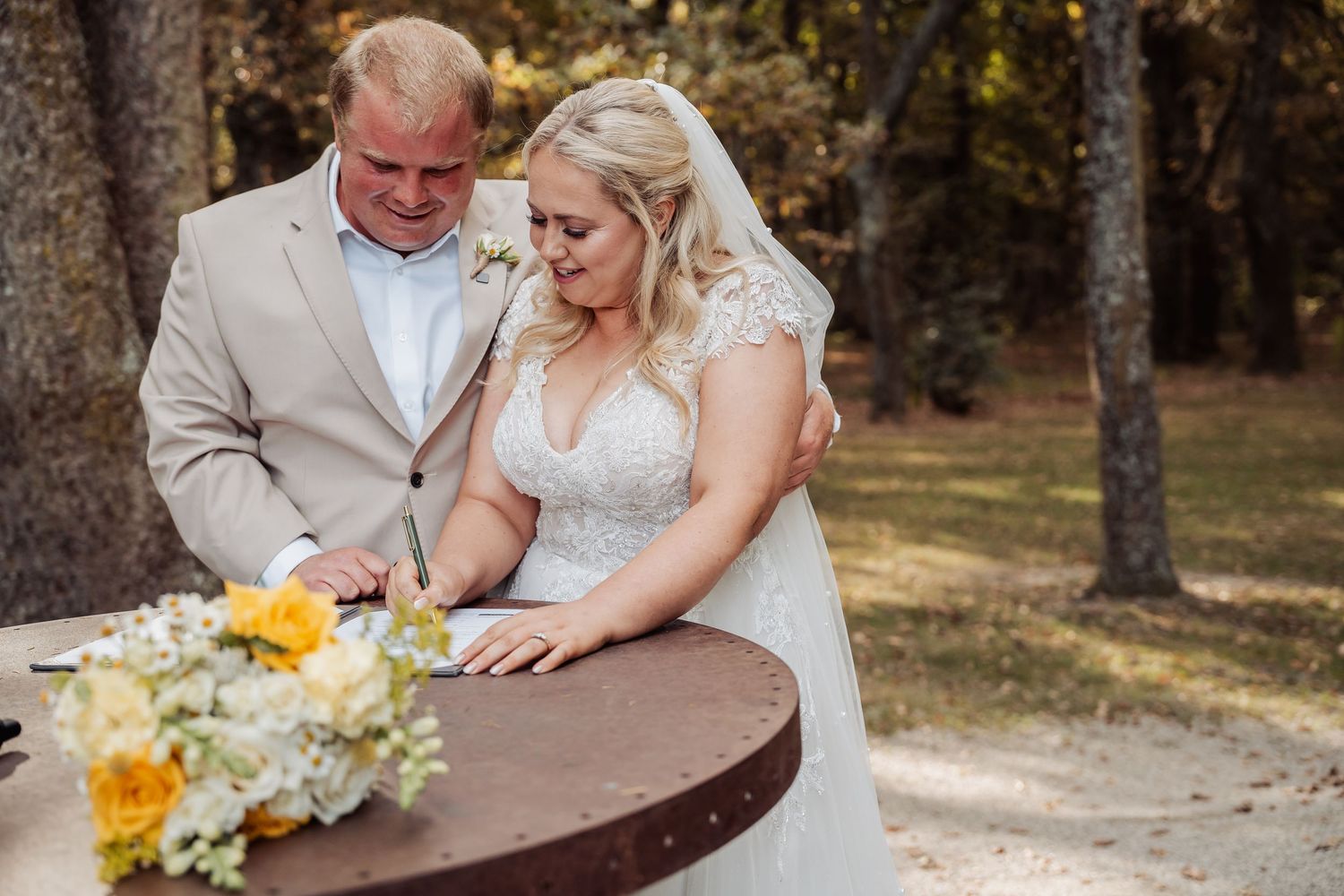 Bride and groom sign wedding documents at outdoor table with yellow floral bouquet nearby.