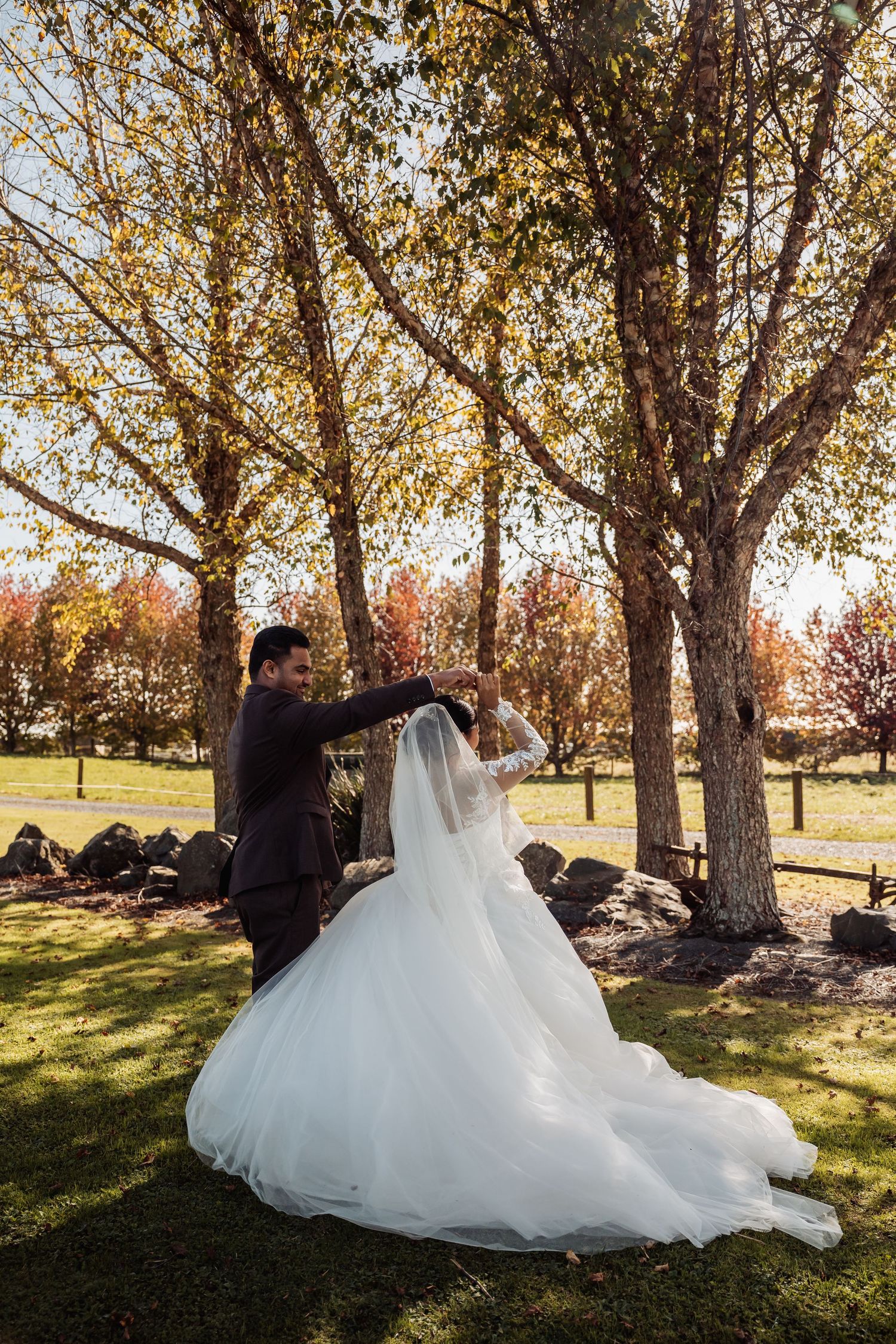 Groom twirling bride in white ball gown outdoors beneath golden autumn trees.