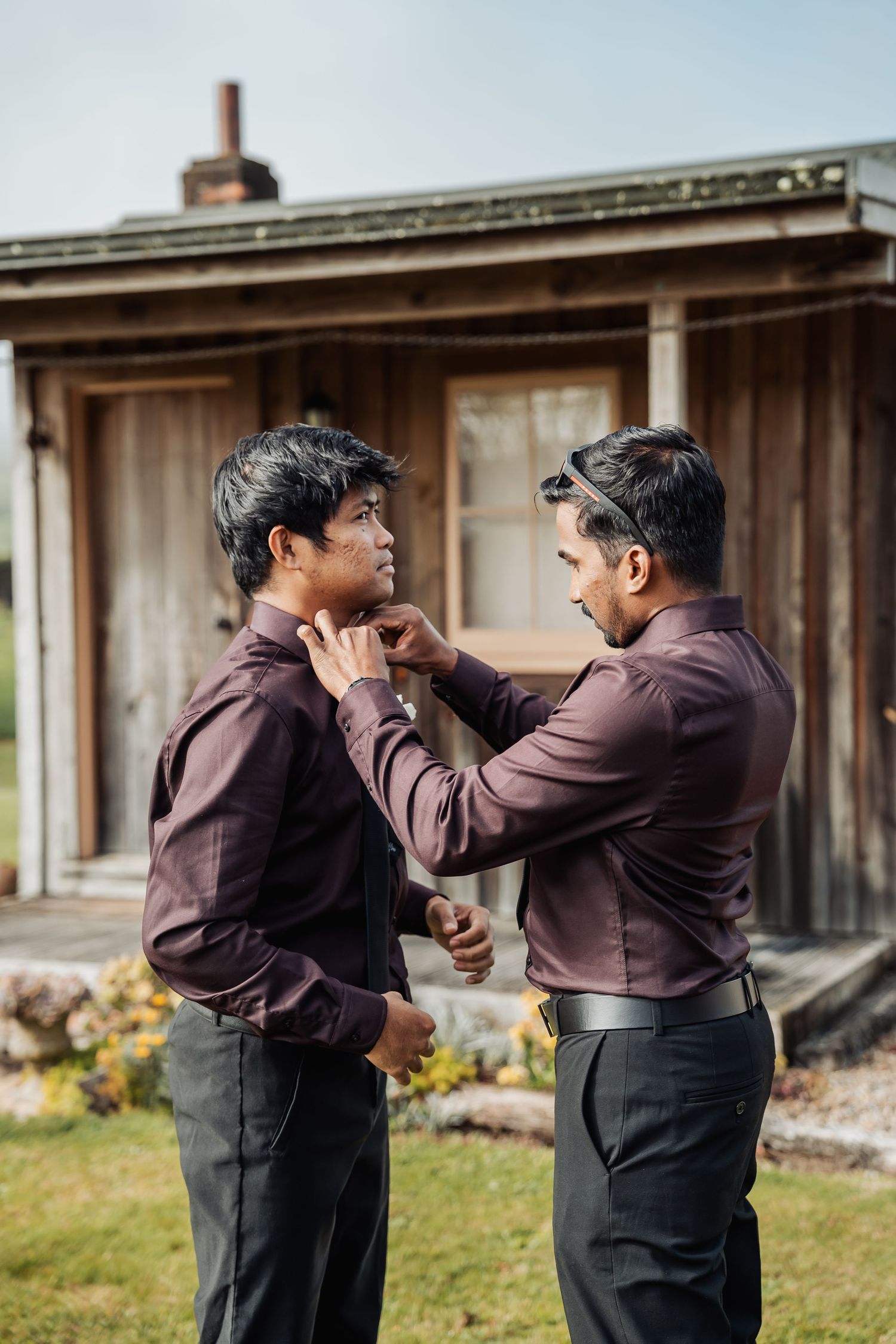 Two groomsmen in matching burgundy shirts help each other prepare outdoors before a wedding ceremony.