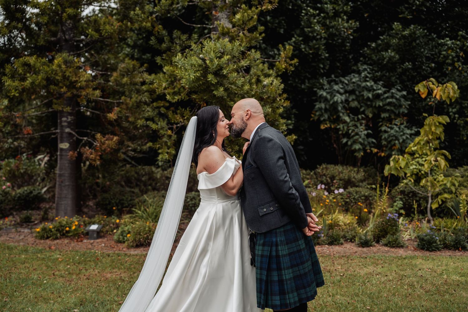 Bride and groom sharing a kiss in a lush garden, bride in white off-shoulder gown, groom in traditional Scottish kilt.