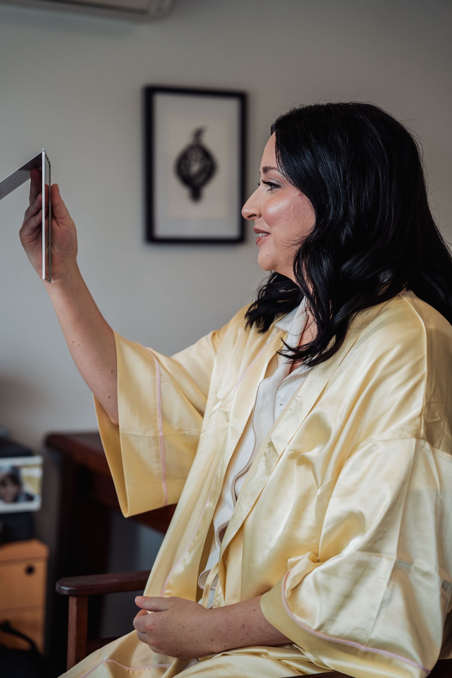 Bride in yellow silk kimono robe holds a mirror while getting ready on her wedding day.