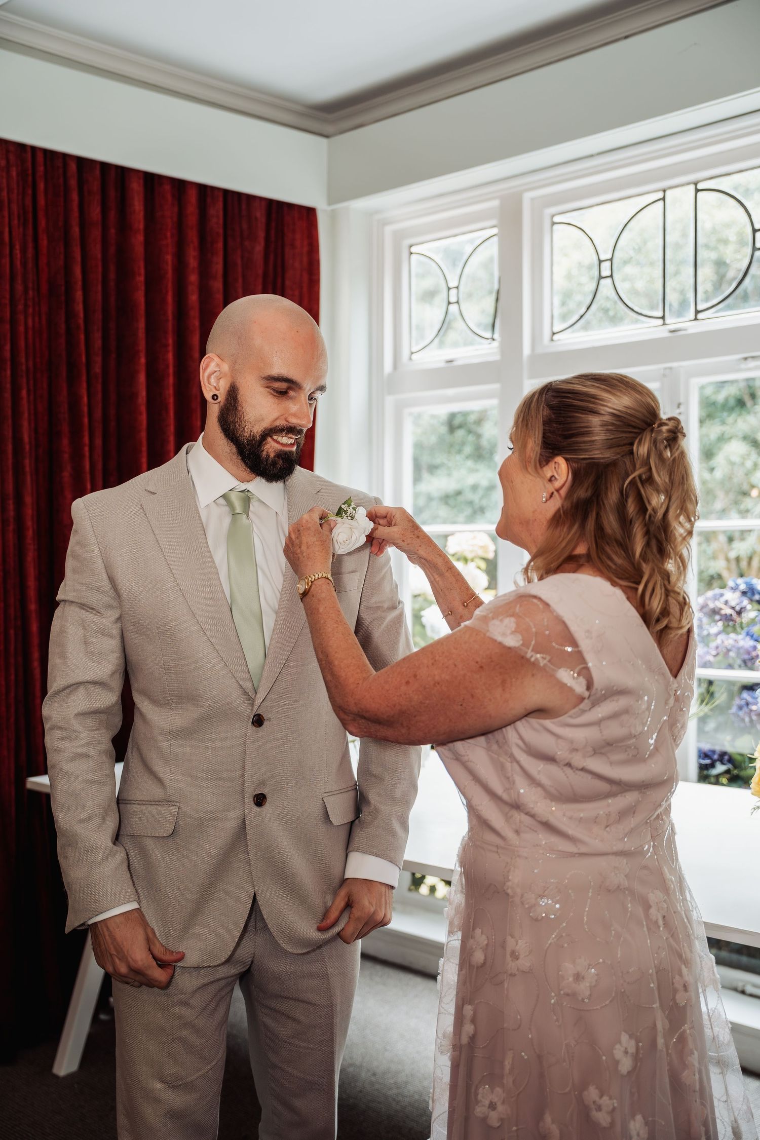 A woman pins a boutonniere on a smiling bald groom in a beige suit with green tie before his wedding.