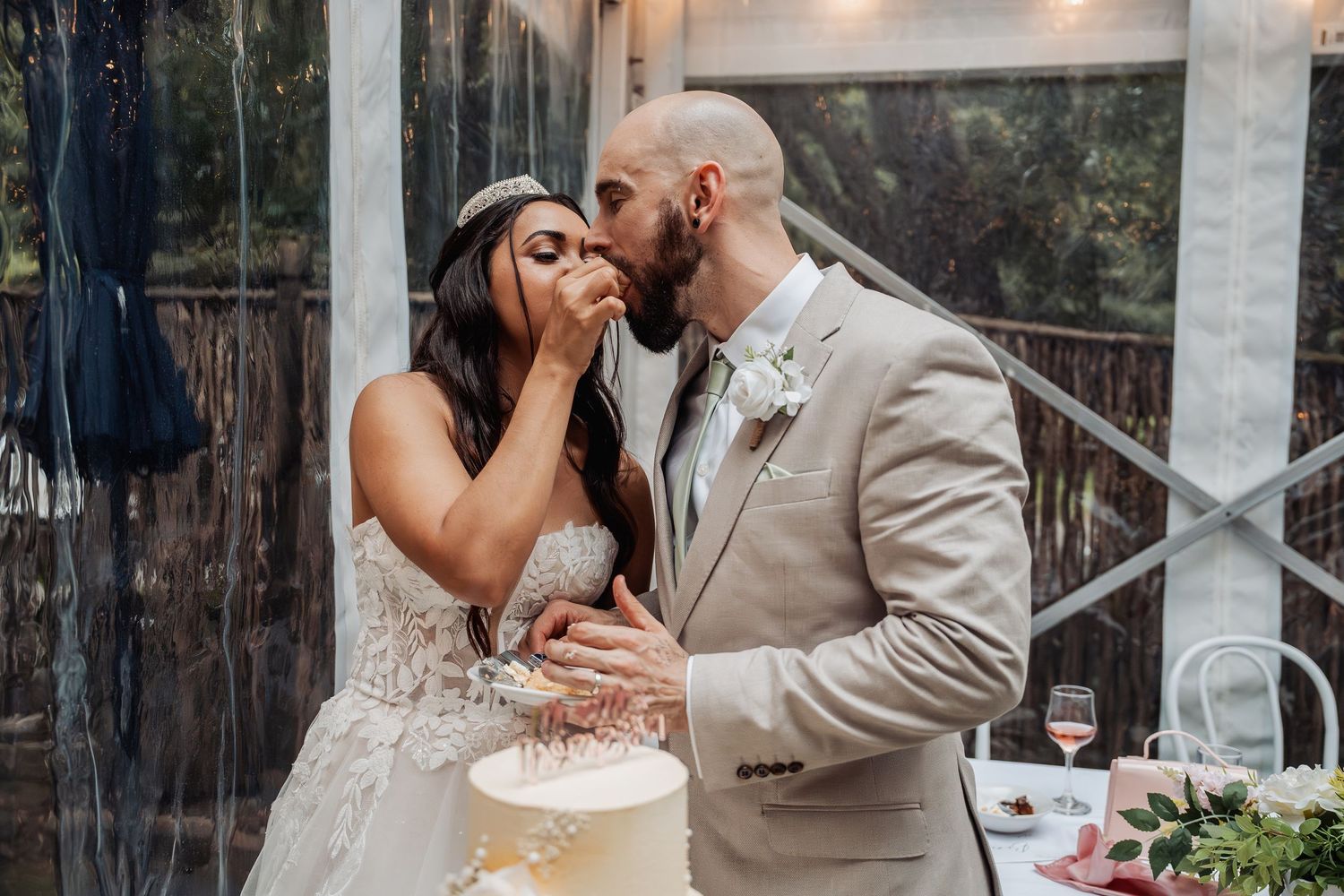 Bride and groom share a kiss while cutting their elegant white wedding cake at reception.
