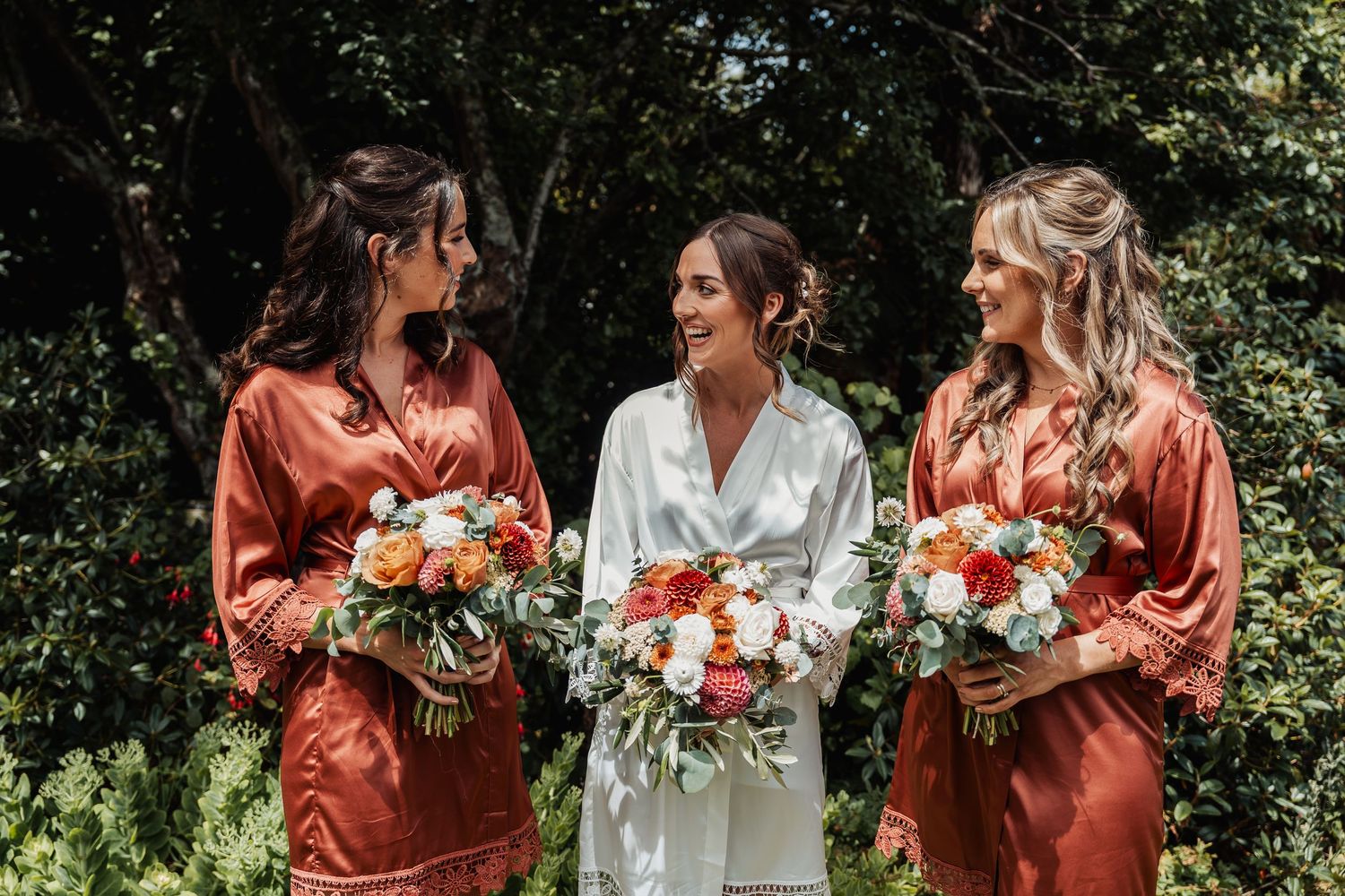 Bride in white robe smiles with two bridesmaids in rust satin robes holding orange floral bouquets outdoors.