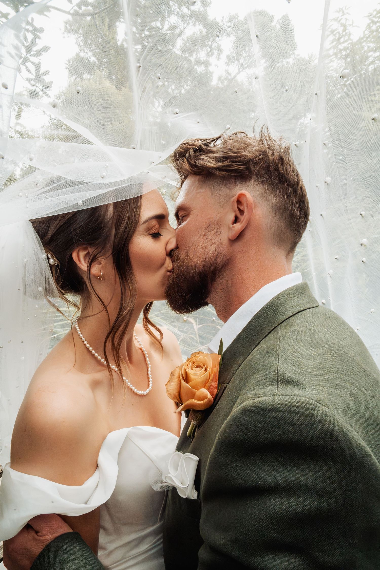 Bride and groom kiss under veil surrounded by lush greenery, groom wearing green suit with rose.