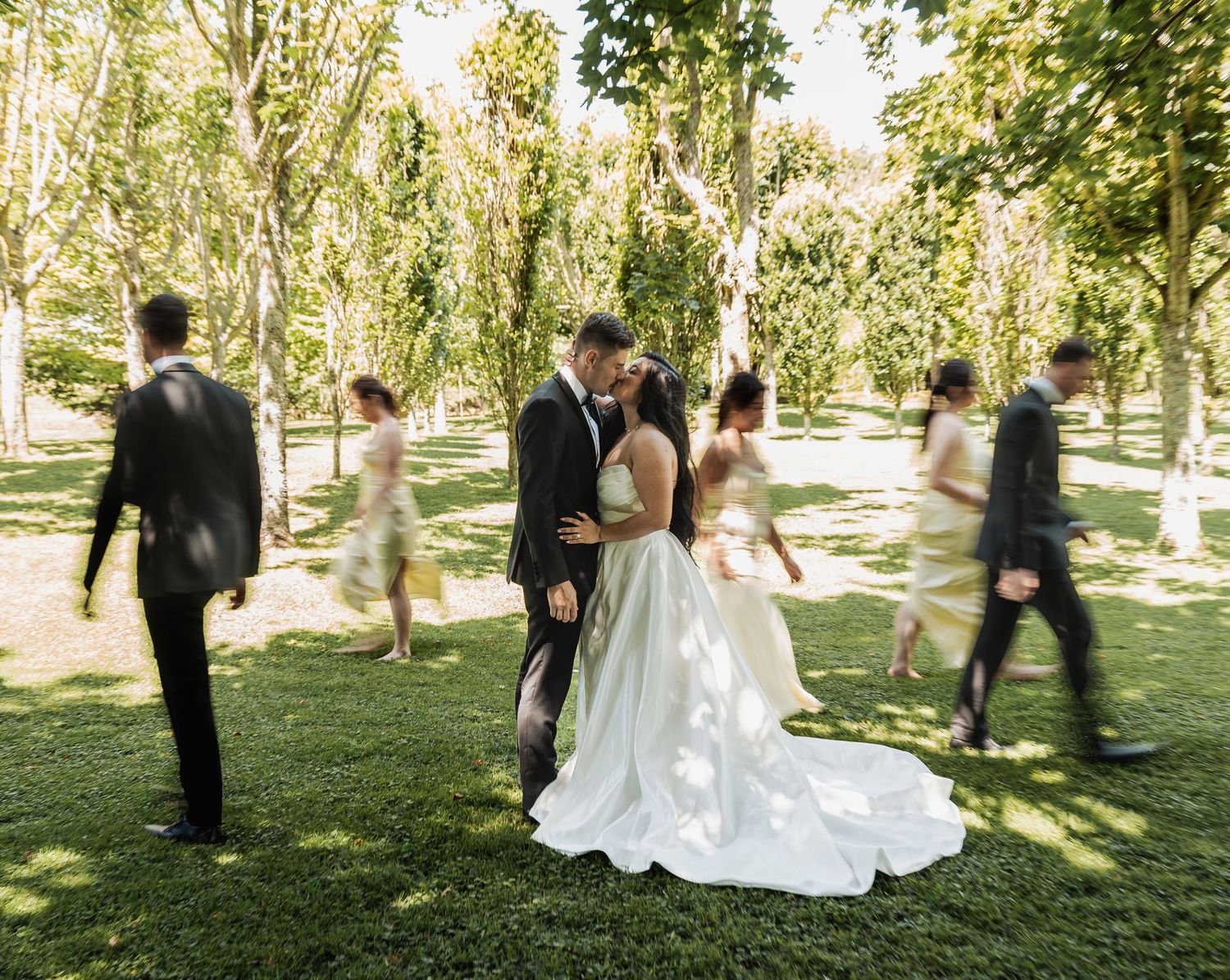 Bride and groom share a kiss in a sunlit garden while wedding party walks around them.