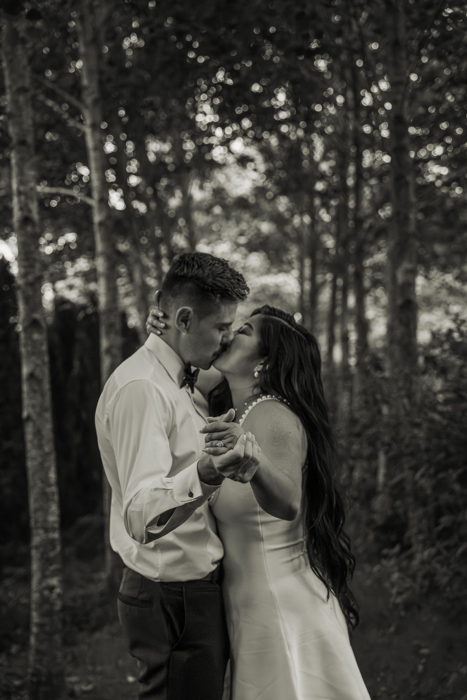 Black and white romantic portrait of bride and groom sharing a kiss among tall trees in forest.