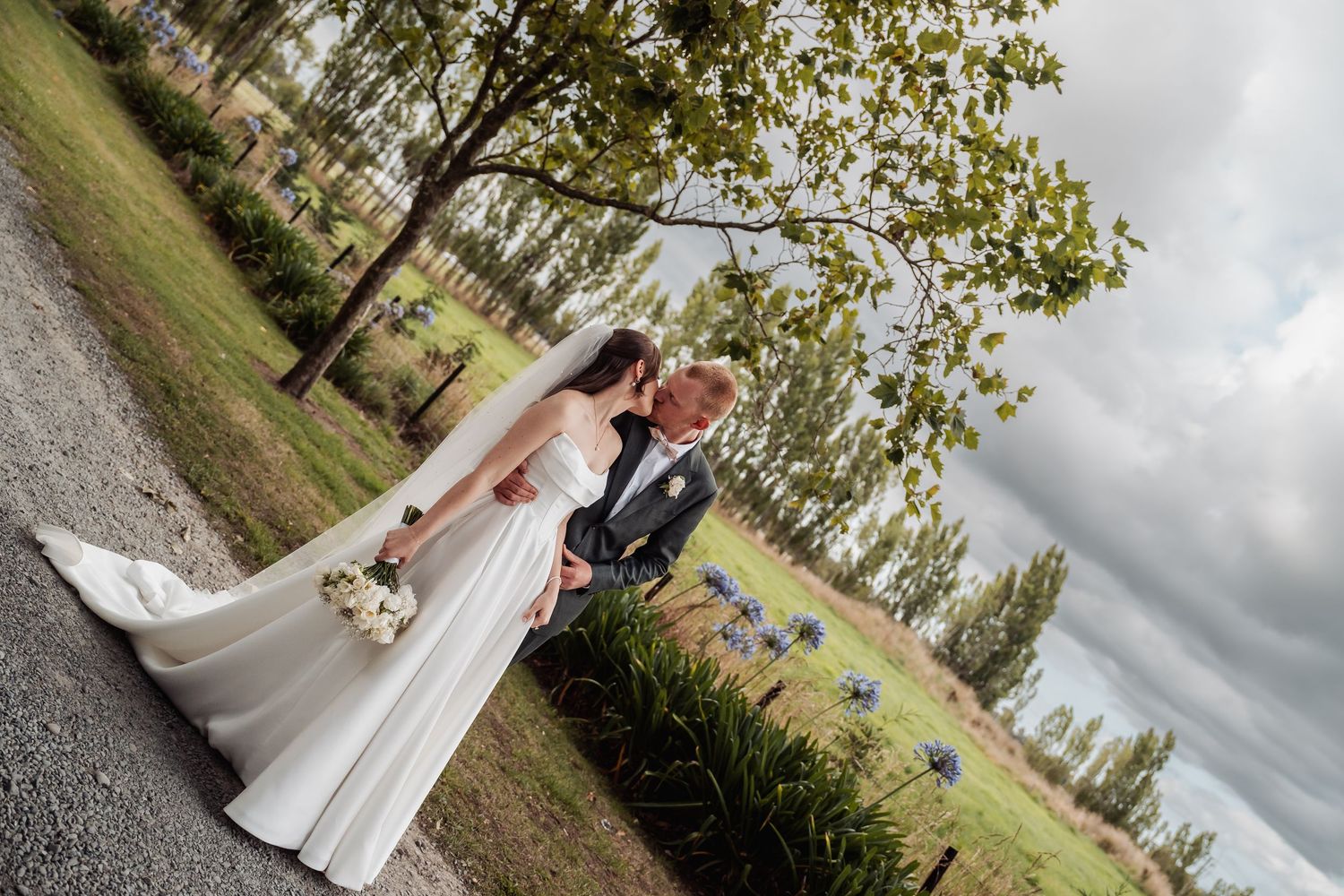 Bride and groom pose romantically outdoors with scenic countryside backdrop featuring tall poplar trees and blue flowers.
