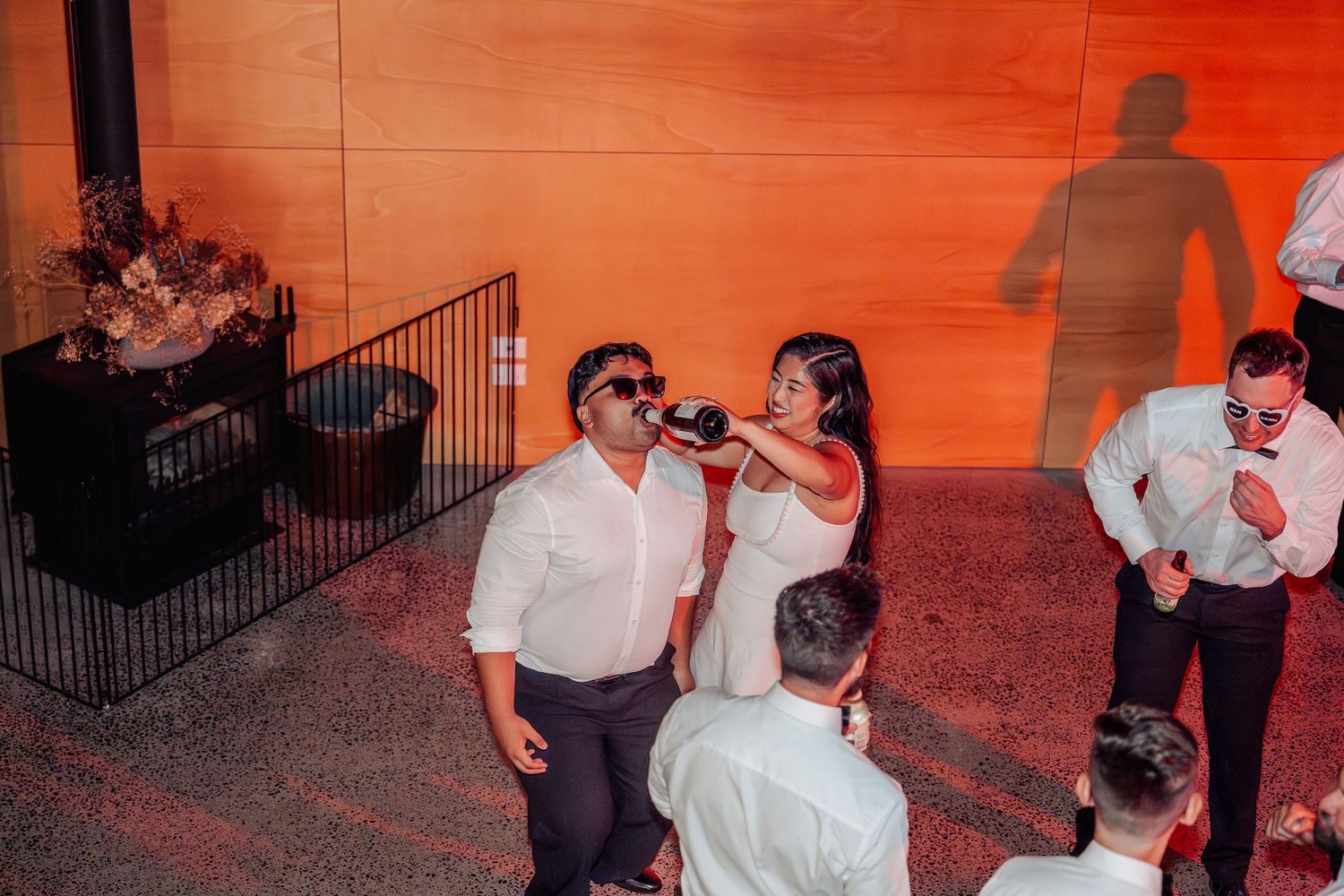 Bride and groom share a playful drink from a bottle wearing sunglasses under warm red reception lighting.