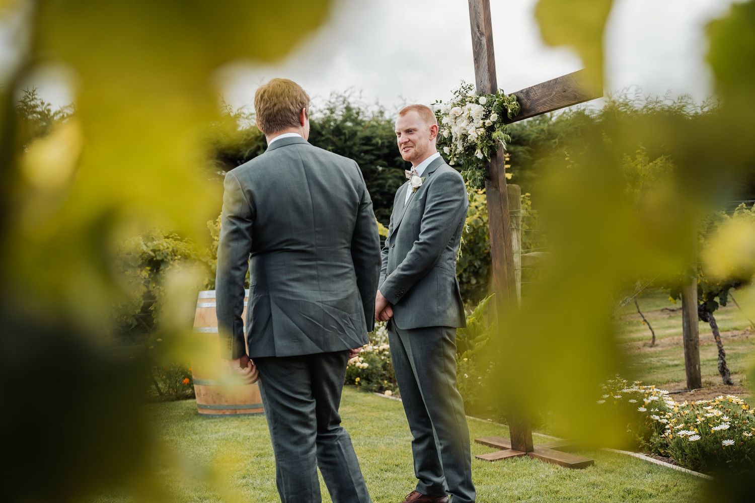 Two groomsmen in grey suits conversing at an outdoor wedding ceremony near a wooden cross altar.