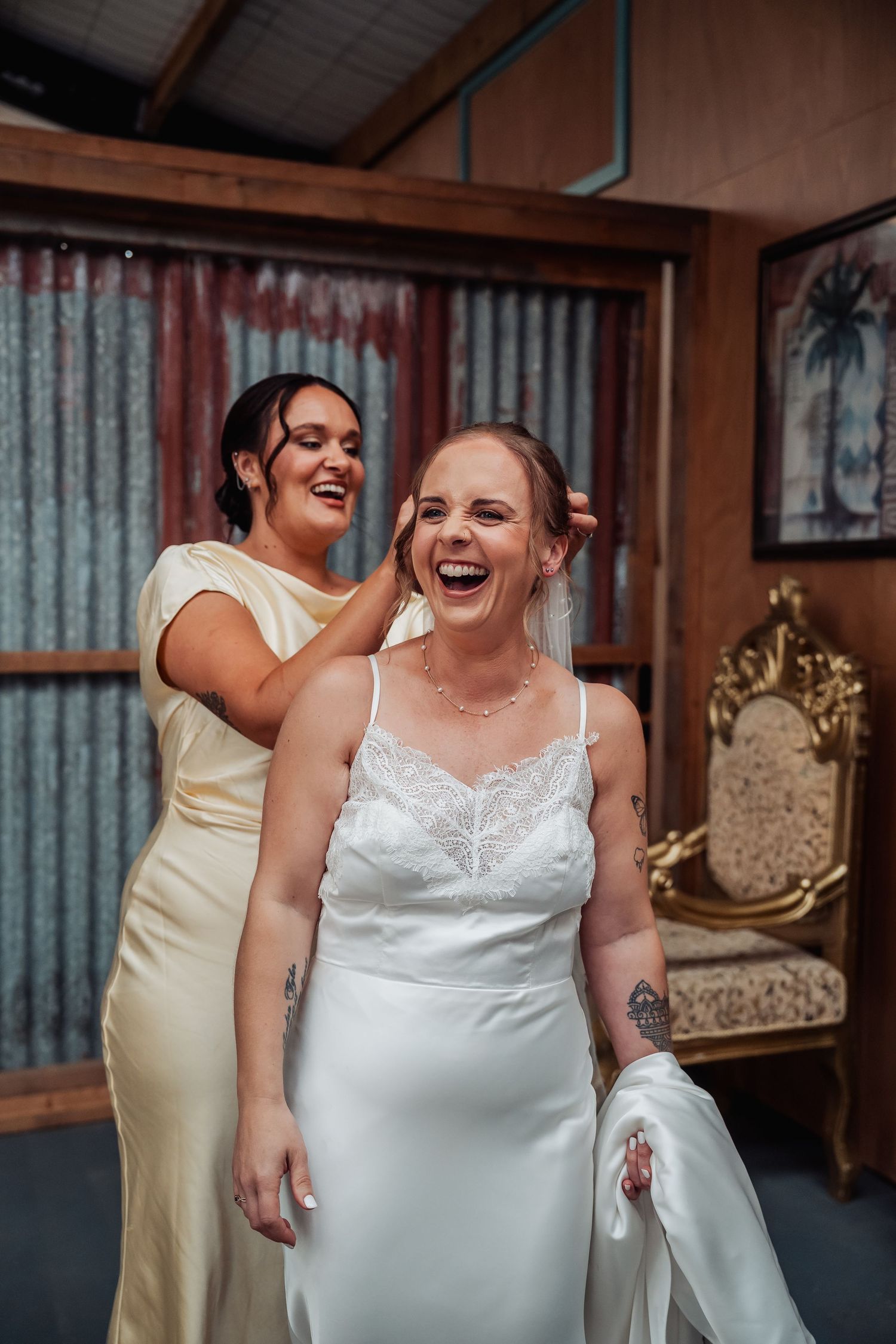 Bride laughing joyfully as bridesmaid adjusts her hair in a rustic wooden room before the wedding ceremony.