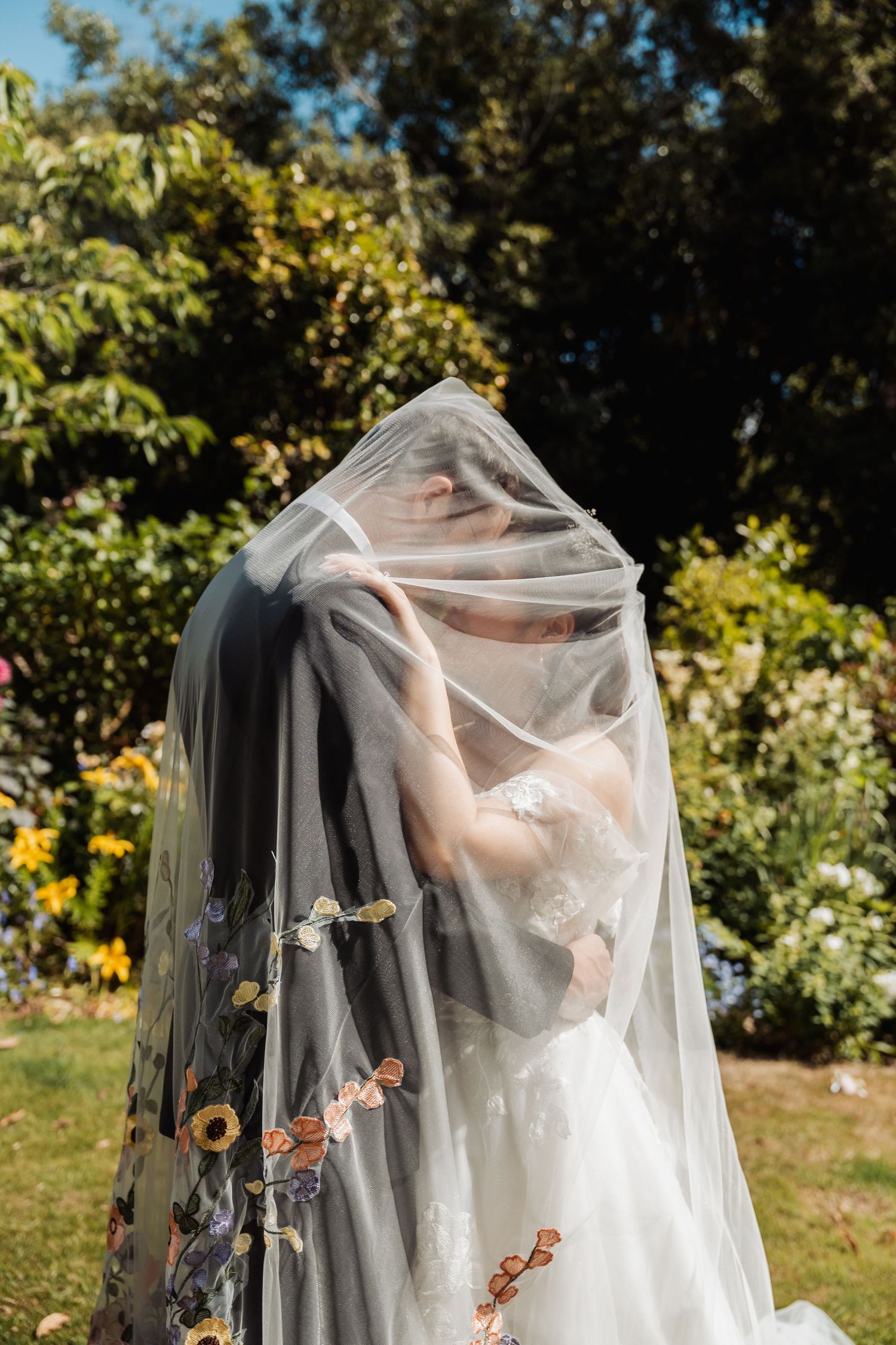 Bride and groom share a kiss hidden beneath a floral embroidered wedding veil outdoors.