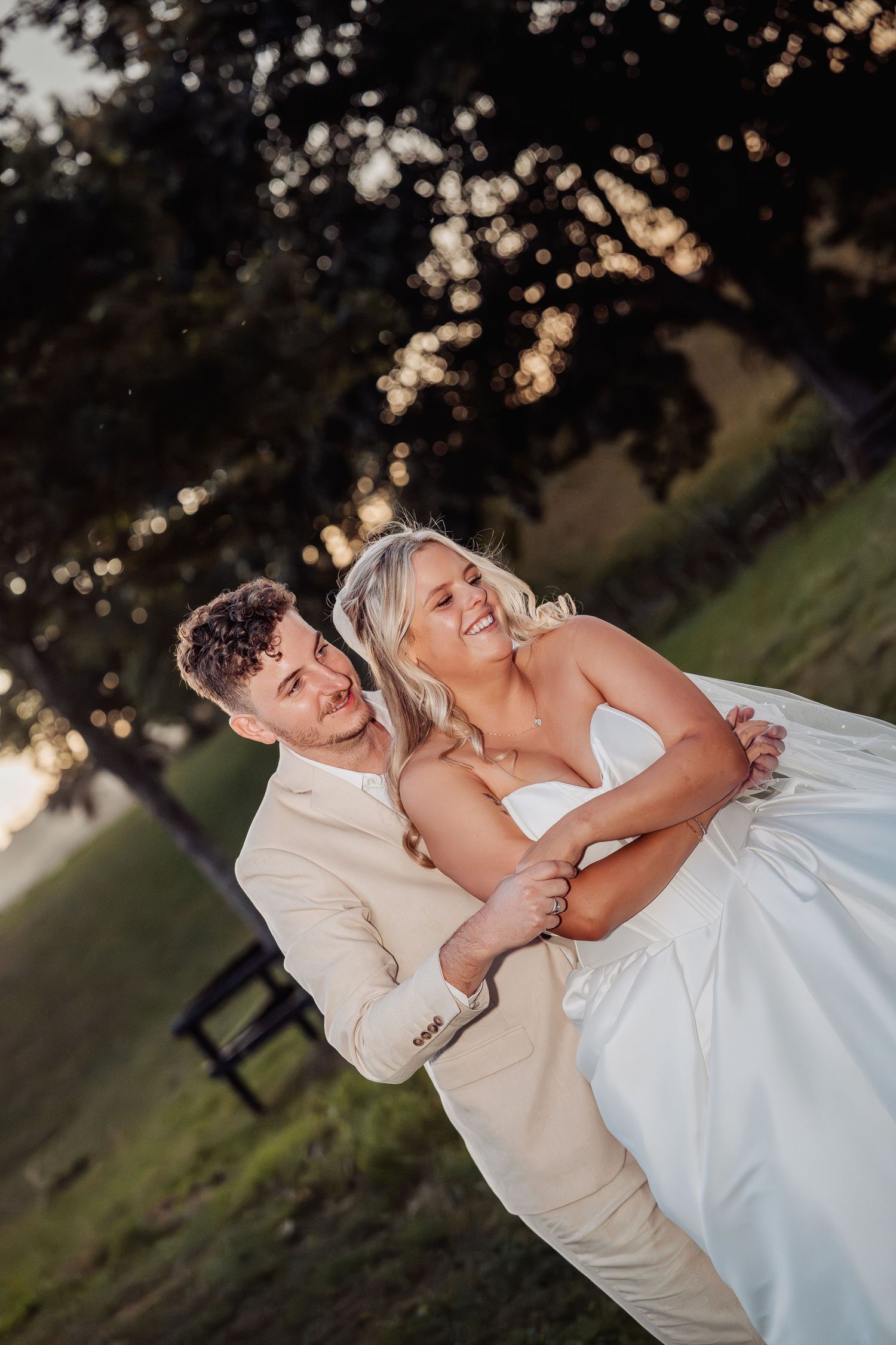 Groom in beige suit joyfully lifting smiling bride in white gown at sunset.