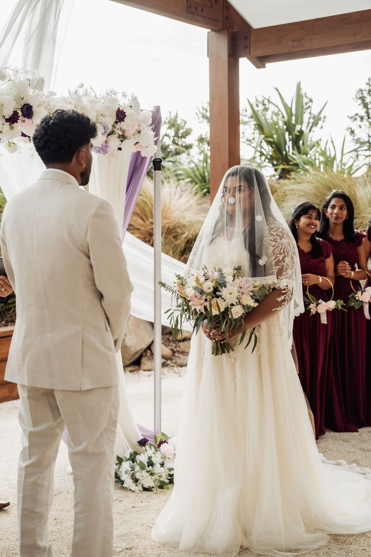 Bride in lace veil holding floral bouquet faces groom at outdoor wedding altar with white flowers.