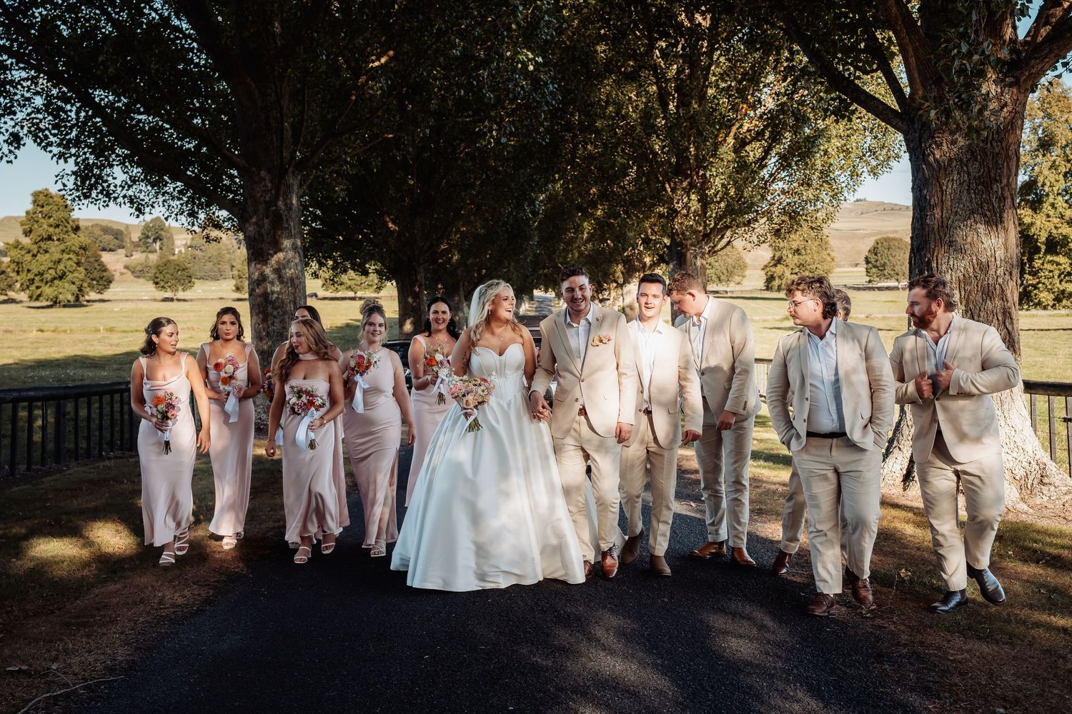 Bride and groom lead wedding party walking together along sun-dappled tree-lined path at outdoor venue.