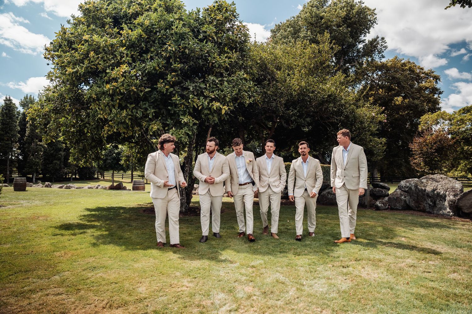 Six groomsmen in matching beige suits walking across a green lawn on a sunny day.