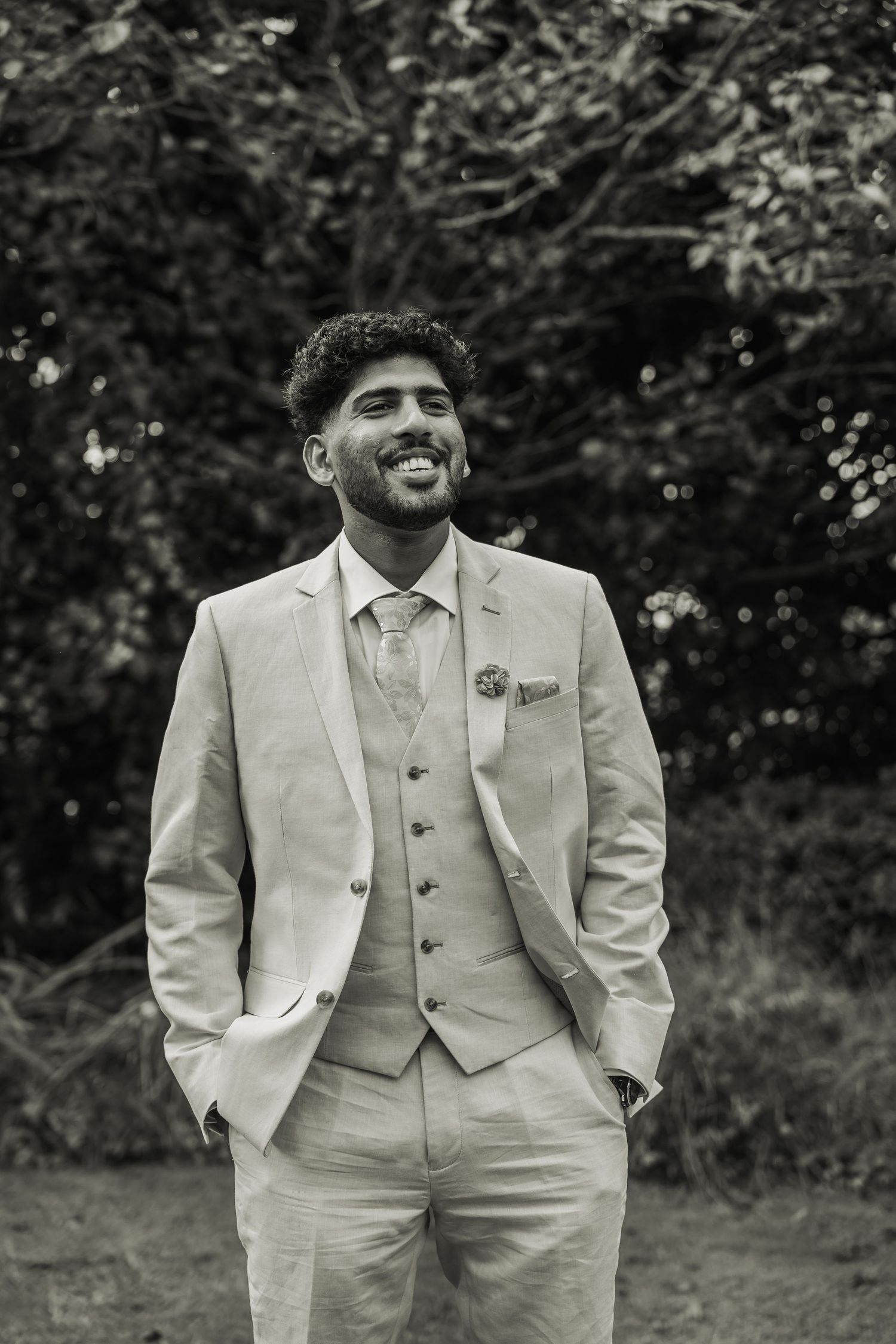Smiling groom in a light beige three-piece suit poses outdoors in a black and white wedding portrait.